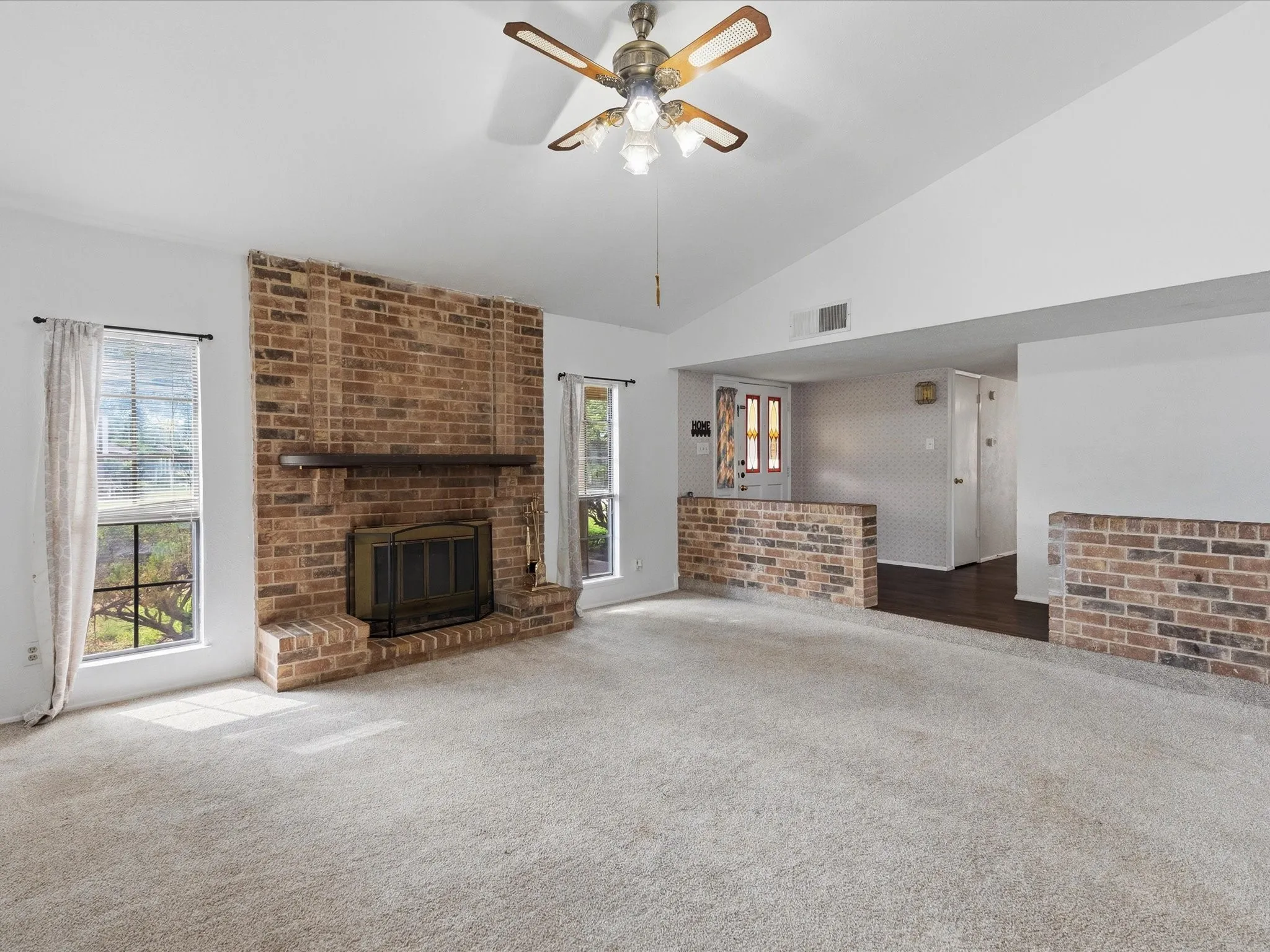 Vaulted ceiling with a ceiling fan, brick fireplace with a wood mantel, light-colored carpeting, and a half-wall brick partition