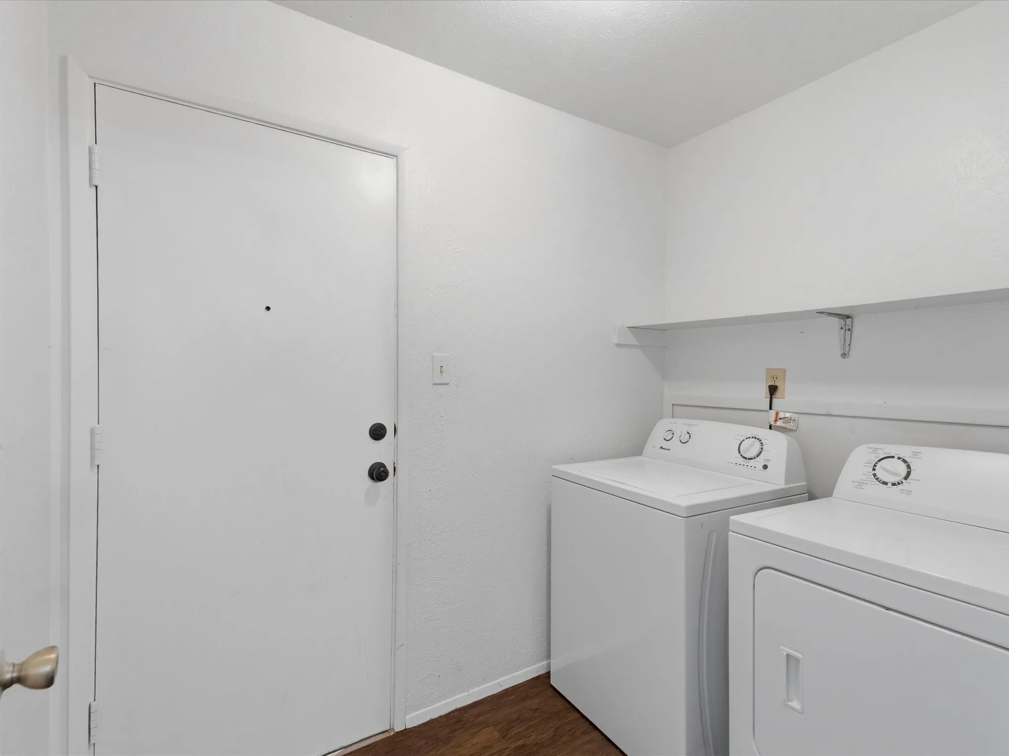 Laundry area with wood-finish flooring, a white shelving unit, and a white entry door with matte black hardware