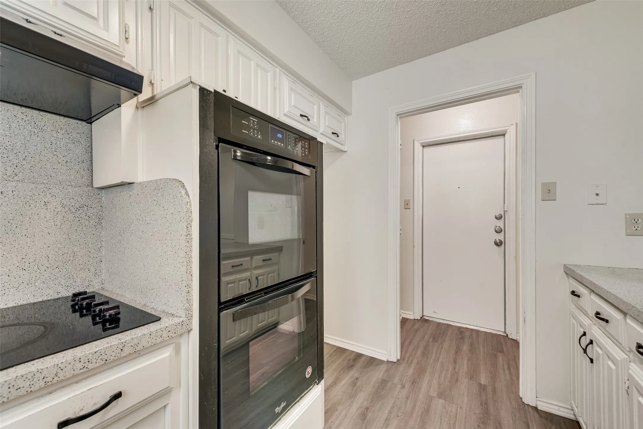 Black built-in double ovens, white cabinetry with dark hardware, a black glass cooktop, light-toned countertops, and wood-finish flooring