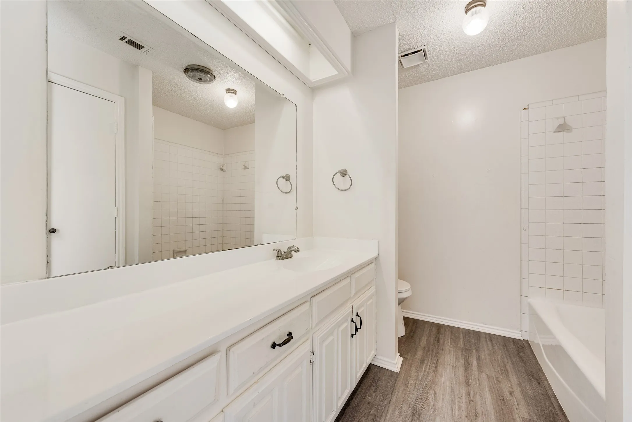 Wood-finish flooring, white vanity with ample storage, large vanity mirror, white tile shower surround, and integrated bathtub