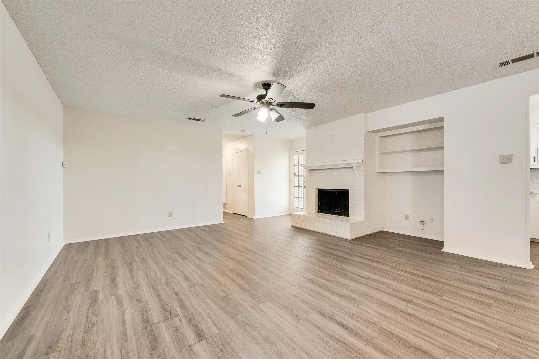 Spacious living area featuring wood-finish flooring, a white brick fireplace, built-in shelving, and a ceiling fan