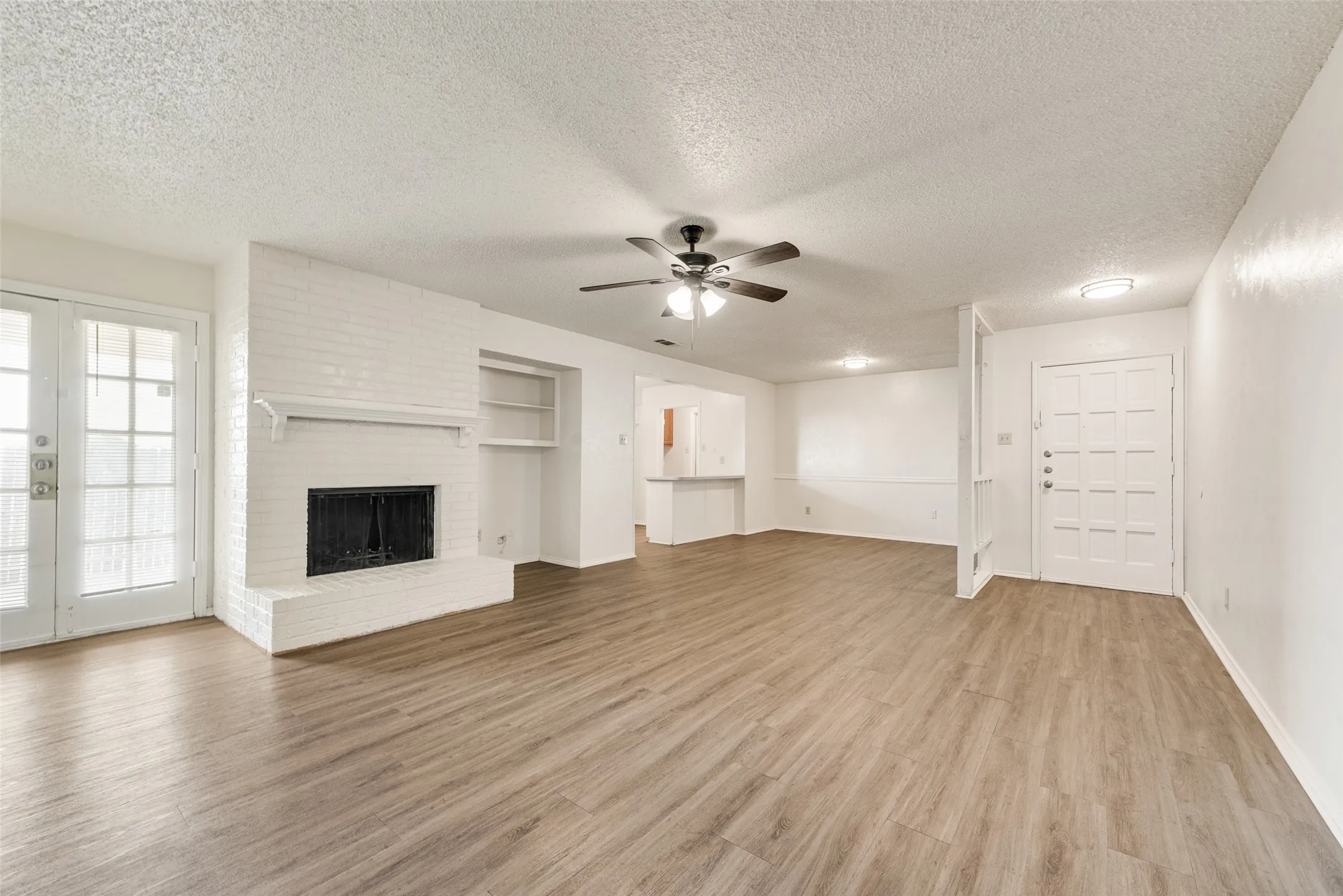 Spacious living area featuring wood-finish flooring, a white brick fireplace with a mantel, built-in shelving, and double French doors