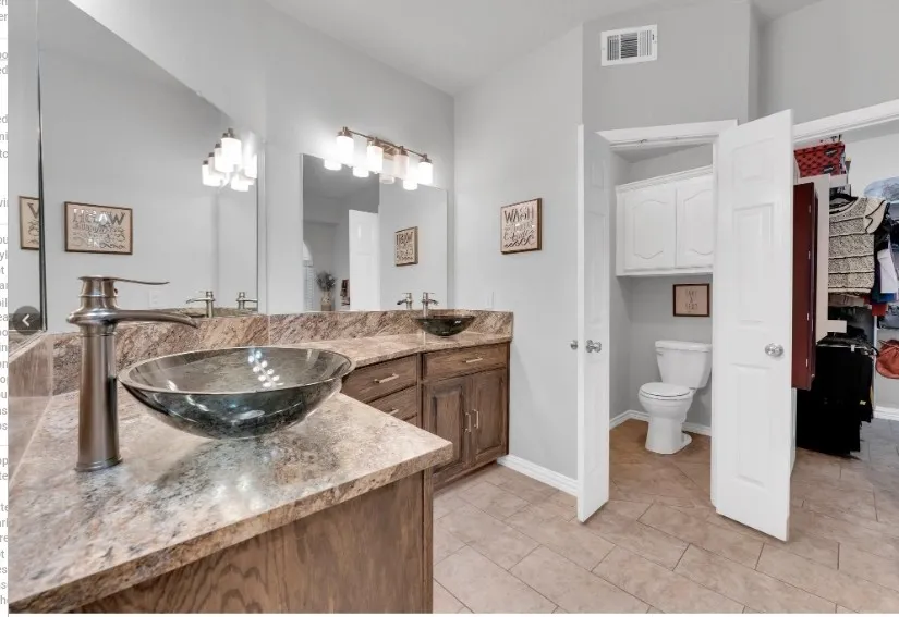 Dual vanity with vessel sinks and bronze-finish faucets, large wall mirror, recessed commode area with built-in cabinetry, and tile flooring