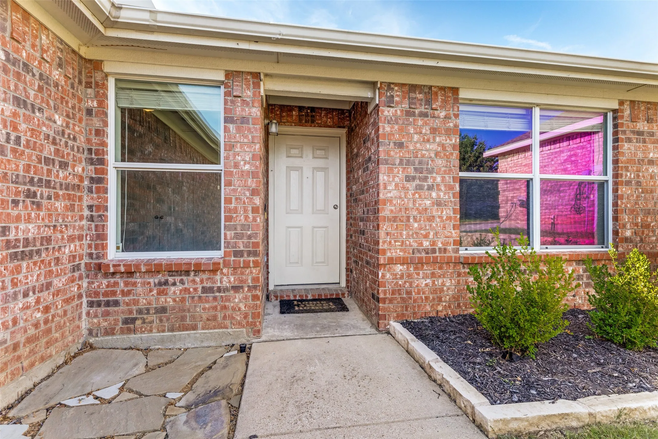 Brick exterior with a paneled entry door, white window frames, concrete pathway, and stone-edged landscaping