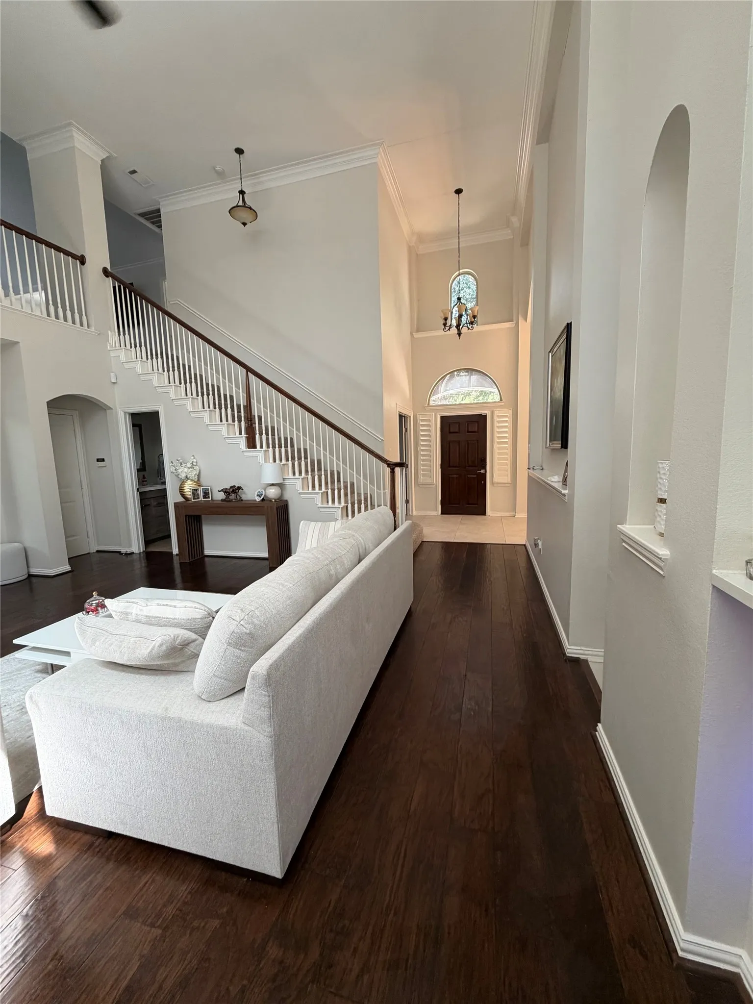 High ceilings with crown molding, dark wood-finish flooring, a grand staircase with white spindles and dark wood handrail, and an arched entryway
