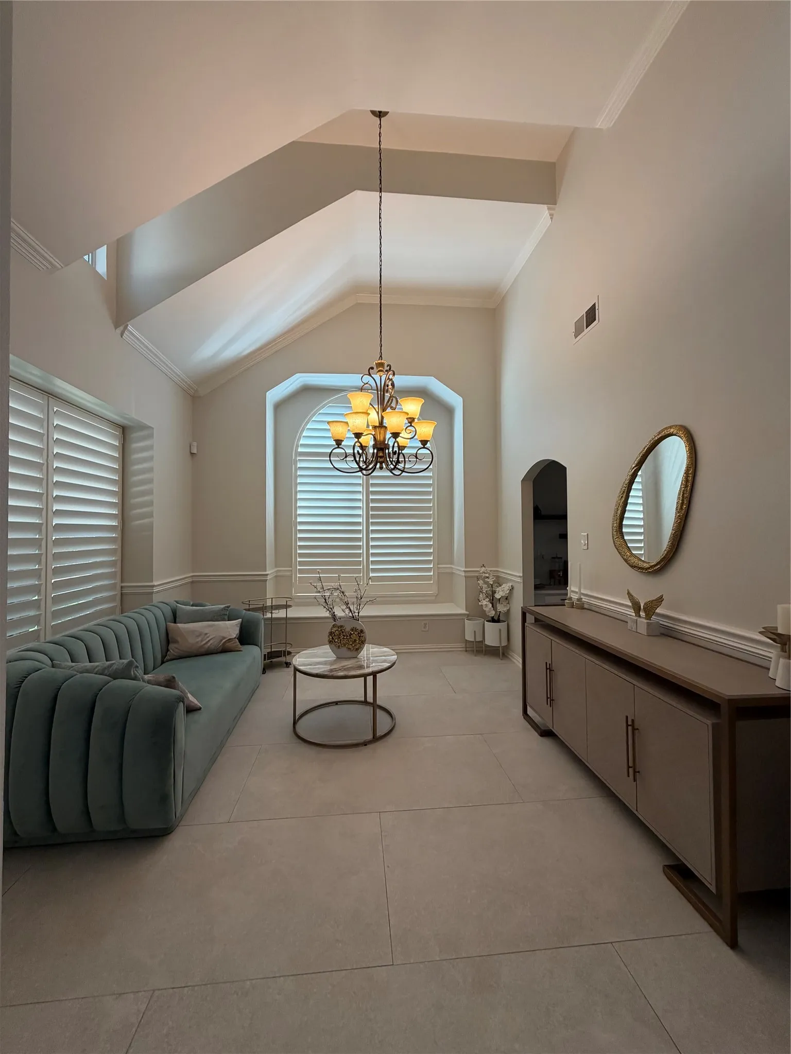 High vaulted ceilings, arched window with plantation shutters, tile flooring, crown molding, and a chandelier