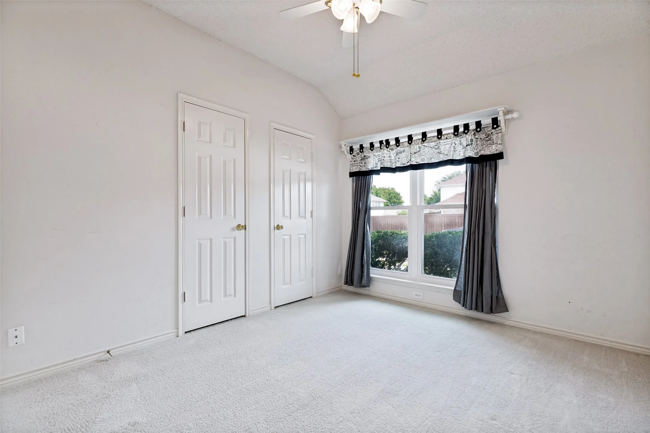 Bright, light-filled bedroom with dual closets featuring six-panel doors, neutral carpet flooring, and large windows dressed with patterned valance and coordinating drapery