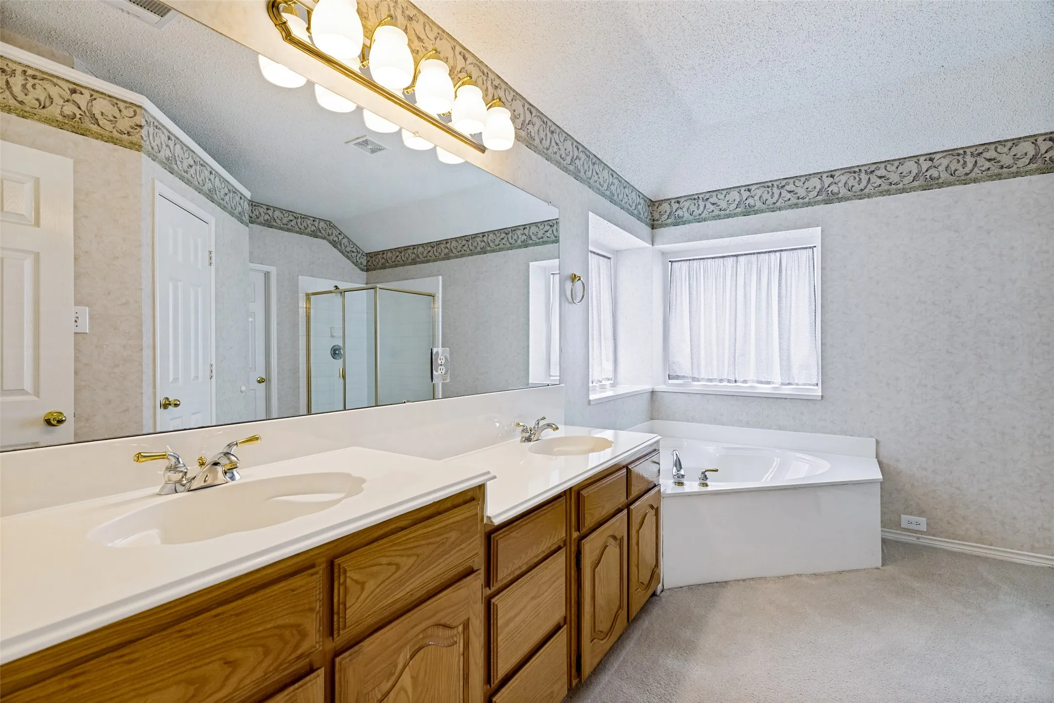 Spa-inspired primary bathroom featuring a dual-sink vanity with wood cabinetry and white quartz countertop, framed mirror with integrated lighting, soaking tub with tile surround, decorative wallpaper border, and abundant natural light from multiple windows