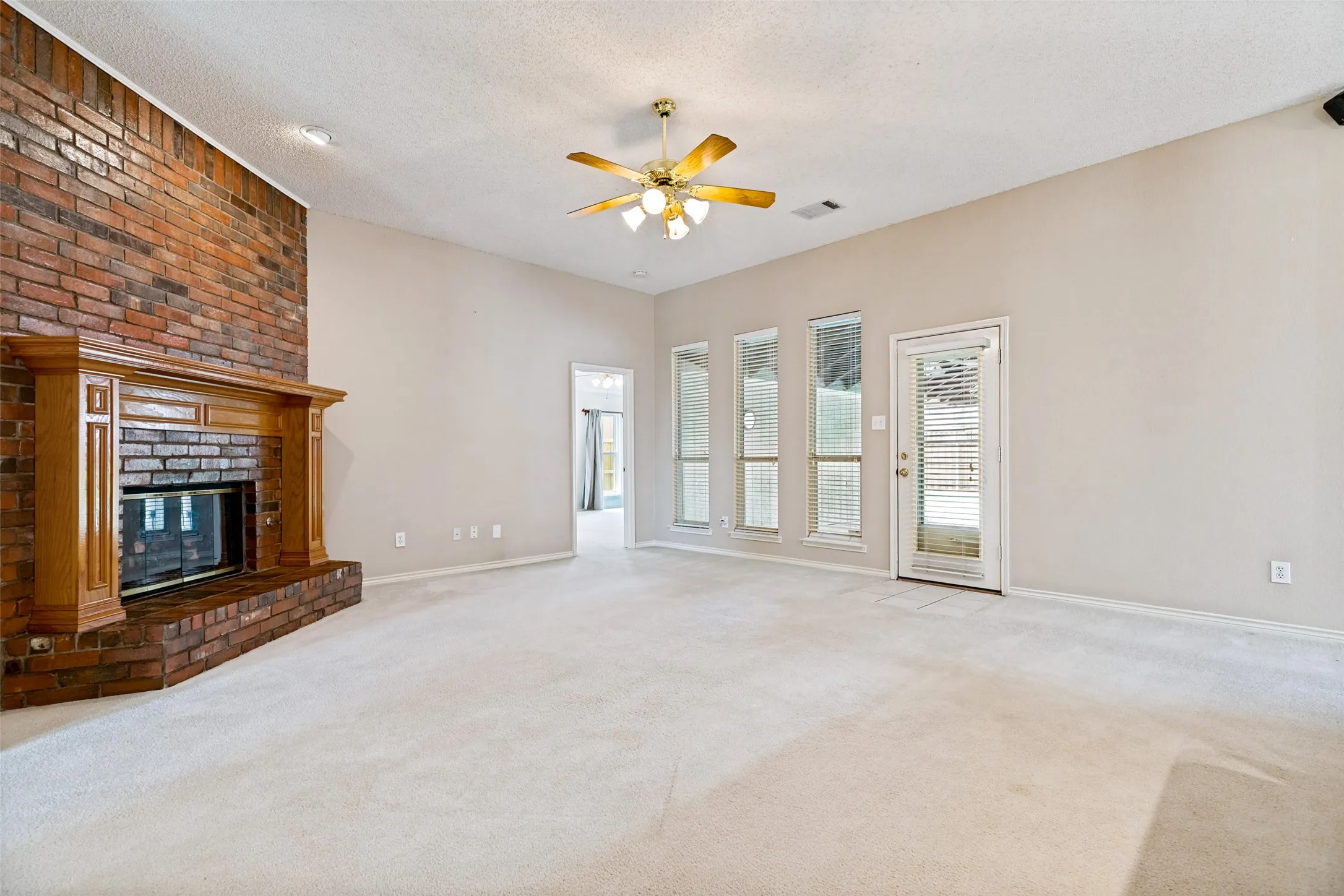 Brick fireplace with wood-finish mantel, vaulted ceiling, ceiling fan with integrated lighting, multiple vertical windows, and a glass-paneled door