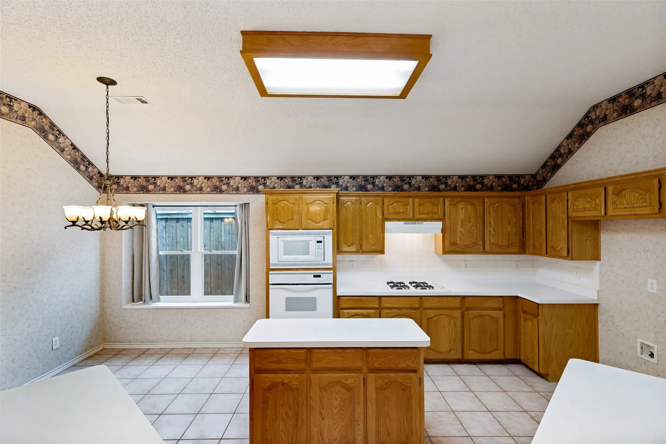 Spacious kitchen featuring warm wood cabinetry, dual work islands with white countertops, ceramic tile flooring, recessed and pendant lighting, and a window overlooking the grounds