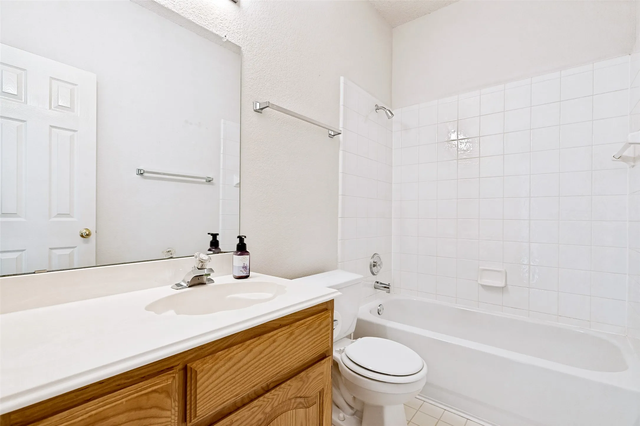 Clean and bright bathroom featuring a wood-finish vanity with ample countertop storage, white subway tile surround, and an integrated bathtub-shower combination with glass enclosure and chrome fixtures