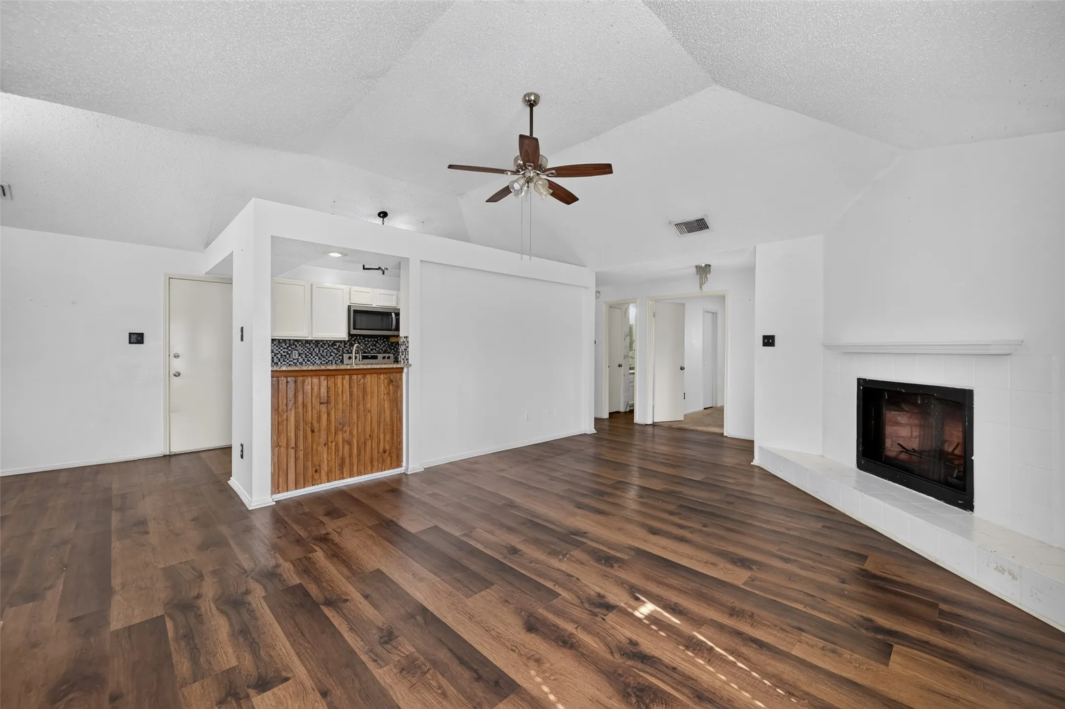 Wood-finish flooring, vaulted ceiling, built-in fireplace, ceiling fan, and a kitchen pass-through with wood-paneled detailing