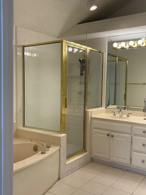 Corner soaking tub, separate shower with brass-finish framing, dual vanity with white cabinetry, and tile flooring