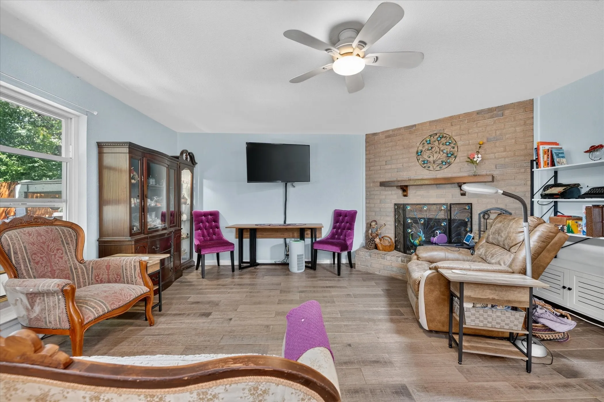 Spacious living area featuring wood-finish flooring, a ceiling fan with integrated lighting, and a brick-faced fireplace with a mantel