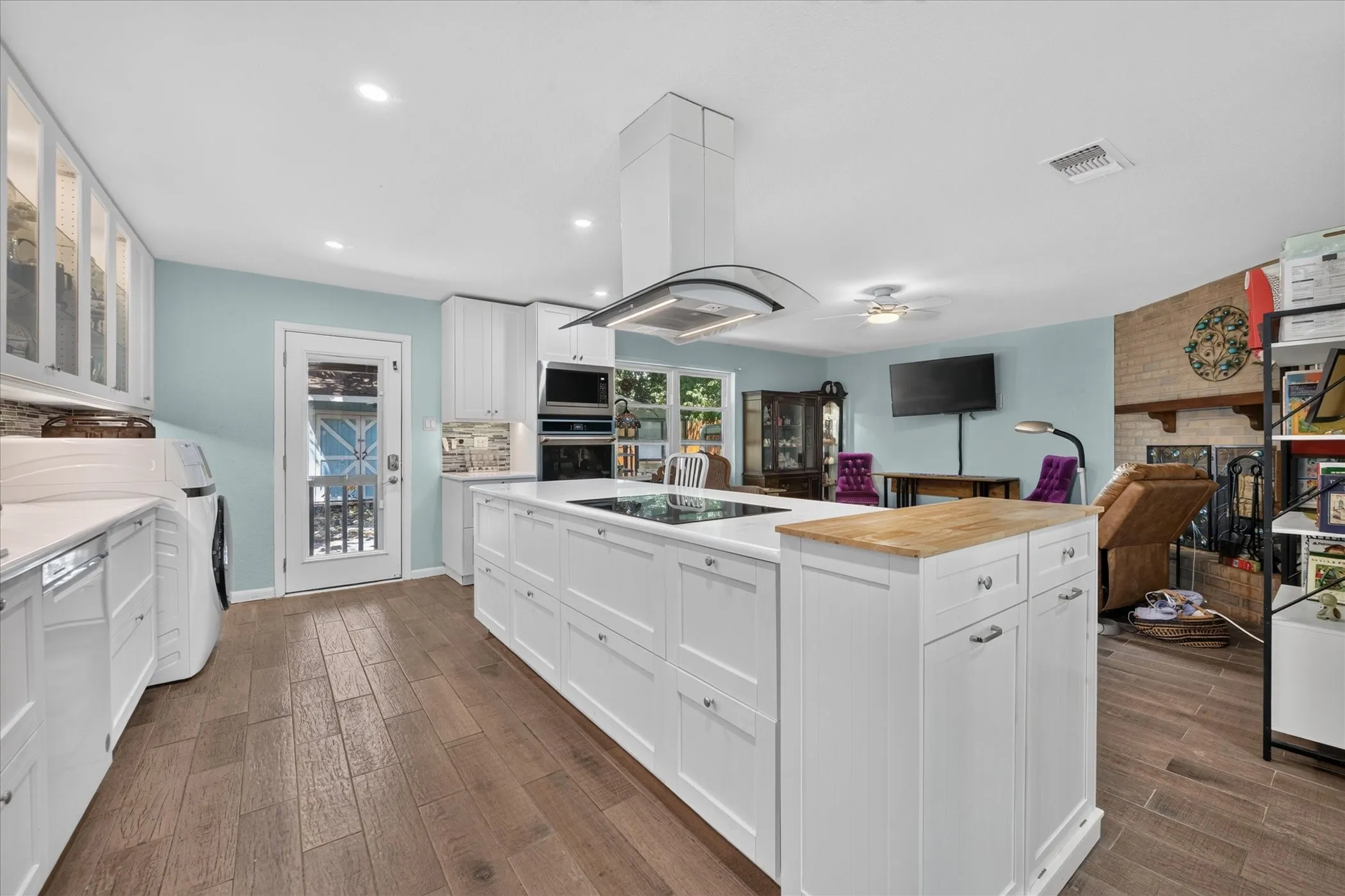 White cabinetry, wood-finish flooring, central island with butcher block and white countertops, built-in stainless steel appliances, and a brick fireplace