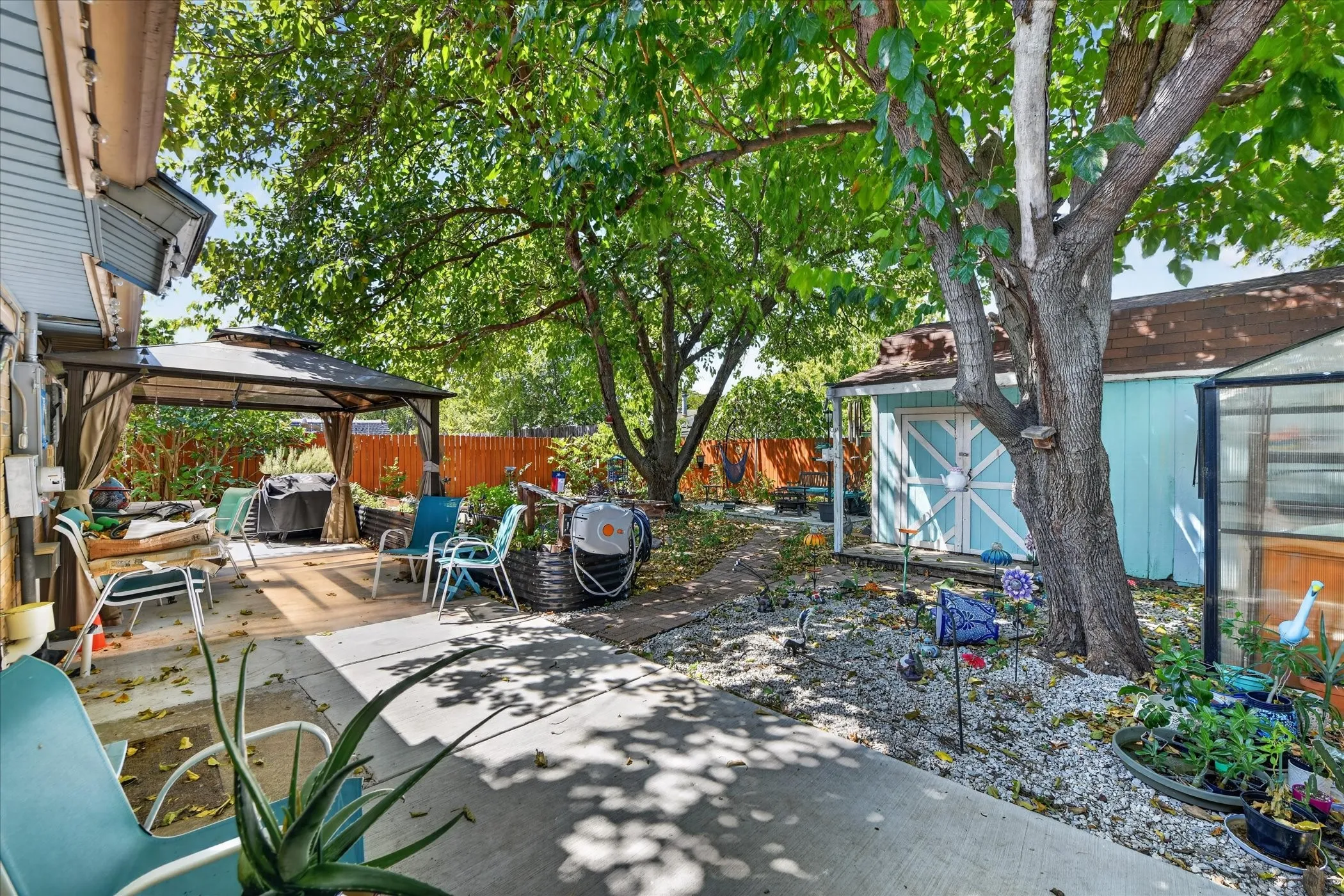 Expansive yard featuring mature trees, a concrete patio, a covered gazebo with a wood-finish deck, a storage shed, and a greenhouse
