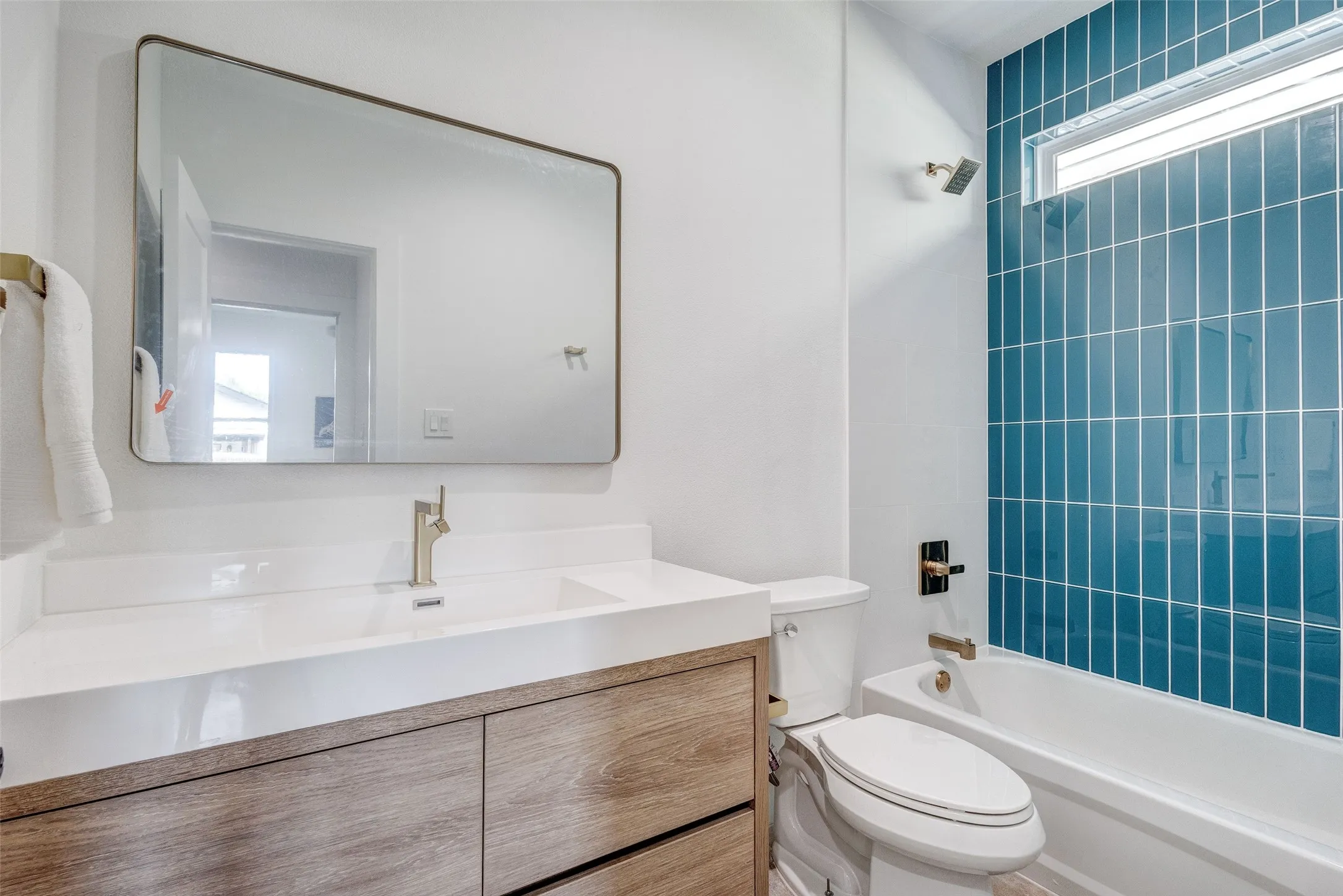Bathroom featuring a wood-finish vanity with an integrated sink, a large framed mirror, a tiled shower-tub combination with a window, and a modern toilet