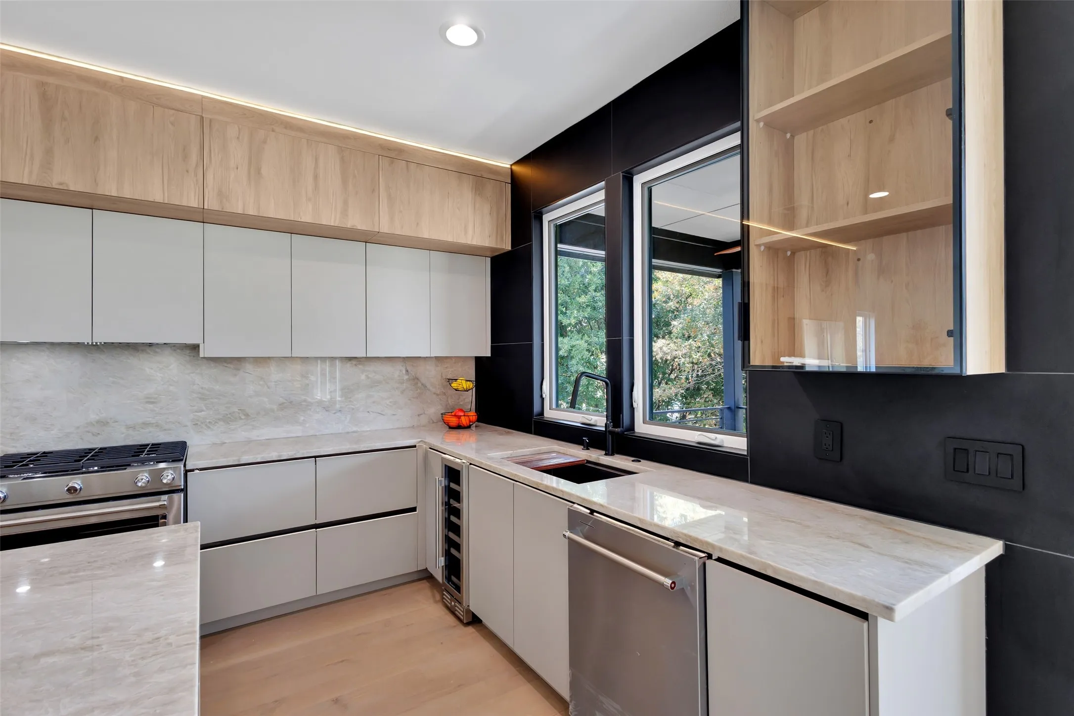 Sleek kitchen featuring light-toned wood-finish cabinetry, a full-height stone backsplash, integrated under-cabinet lighting, and stainless steel appliances