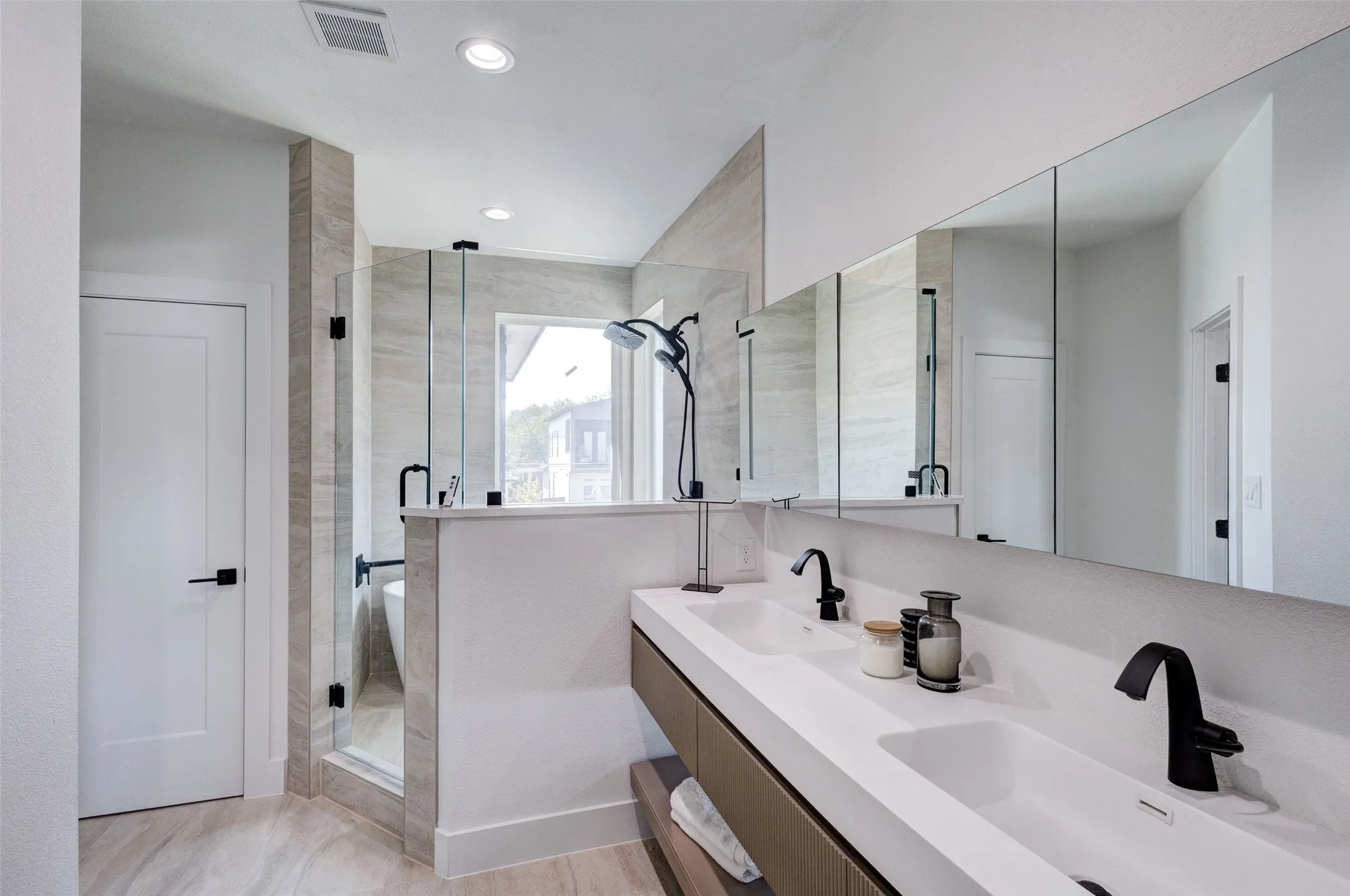 Dual integrated sinks with matte black faucets, floating vanity with wood-finish cabinetry, frameless mirrored medicine cabinets, glass-enclosed shower with a rainfall showerhead, and a freestanding soaking tub