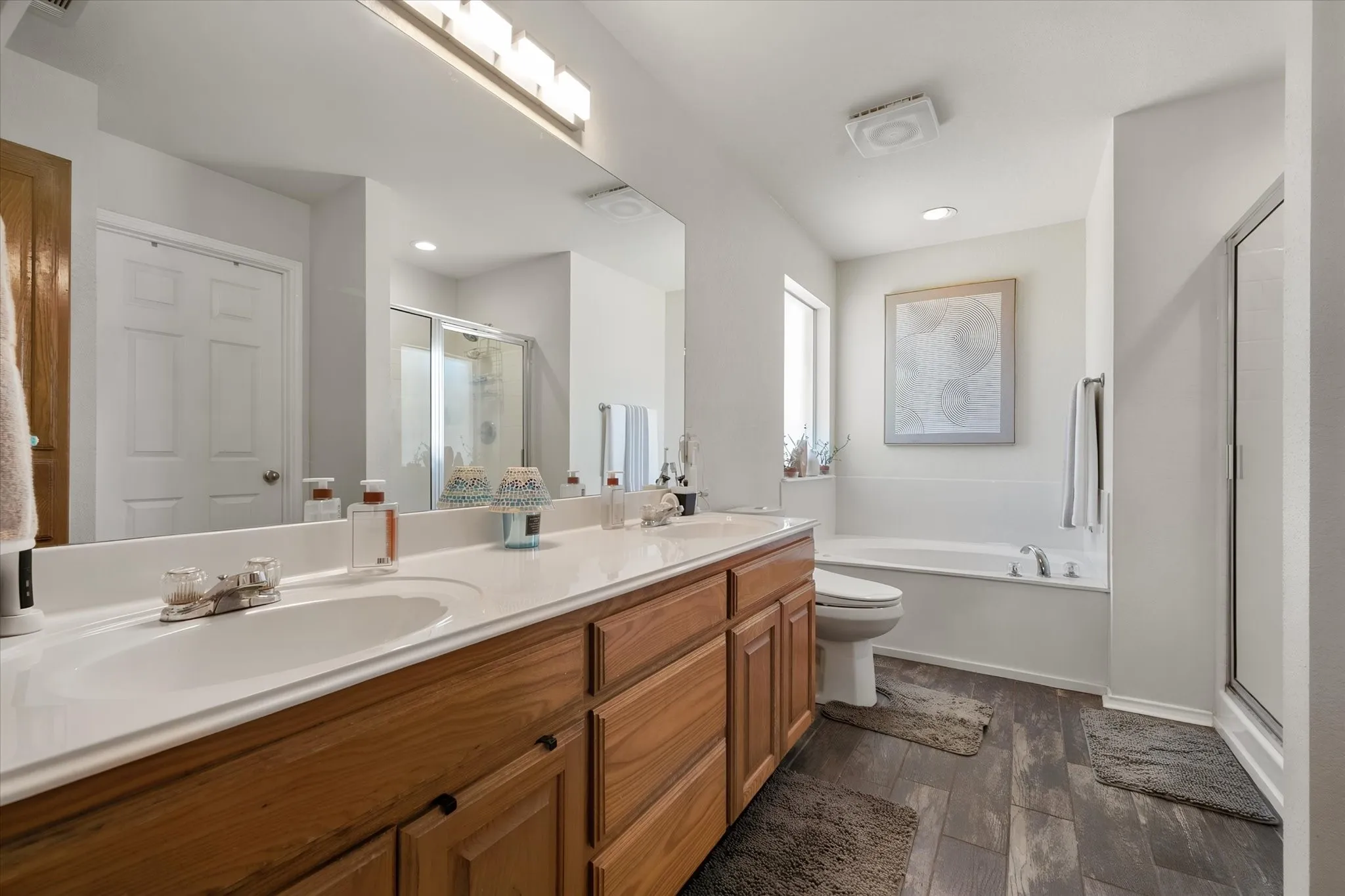 Double vanity with integrated sinks, wood-finish cabinetry, built-in bathtub, glass-enclosed shower, and wood-finish flooring