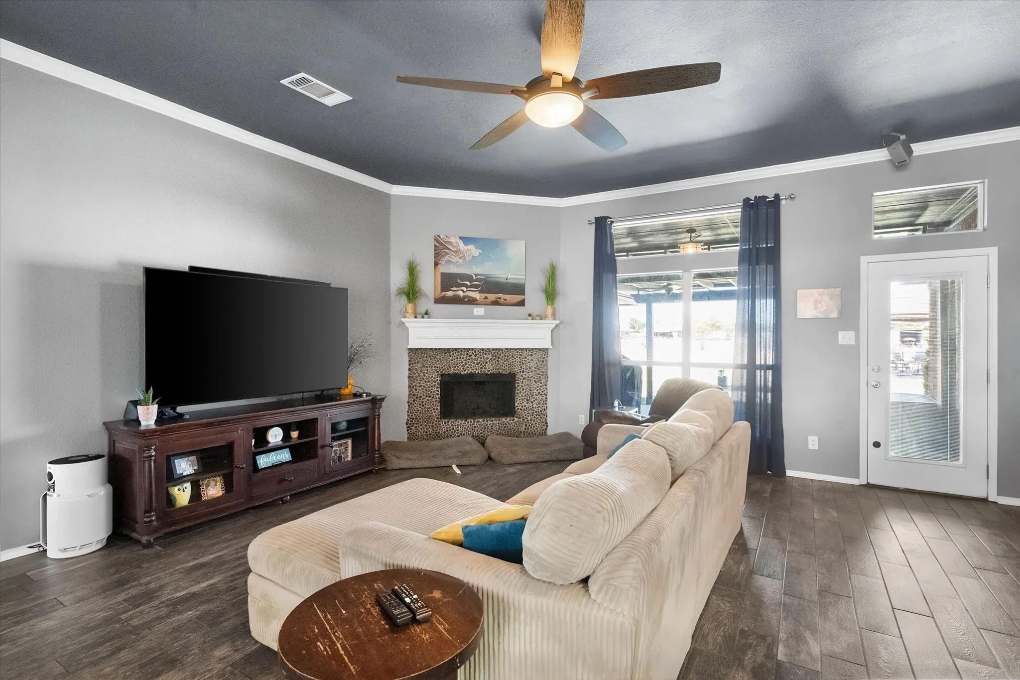 Wood-finish flooring, ceiling fan with integrated lighting, fireplace with stone surround and mantel, white crown molding, and glass-paneled door