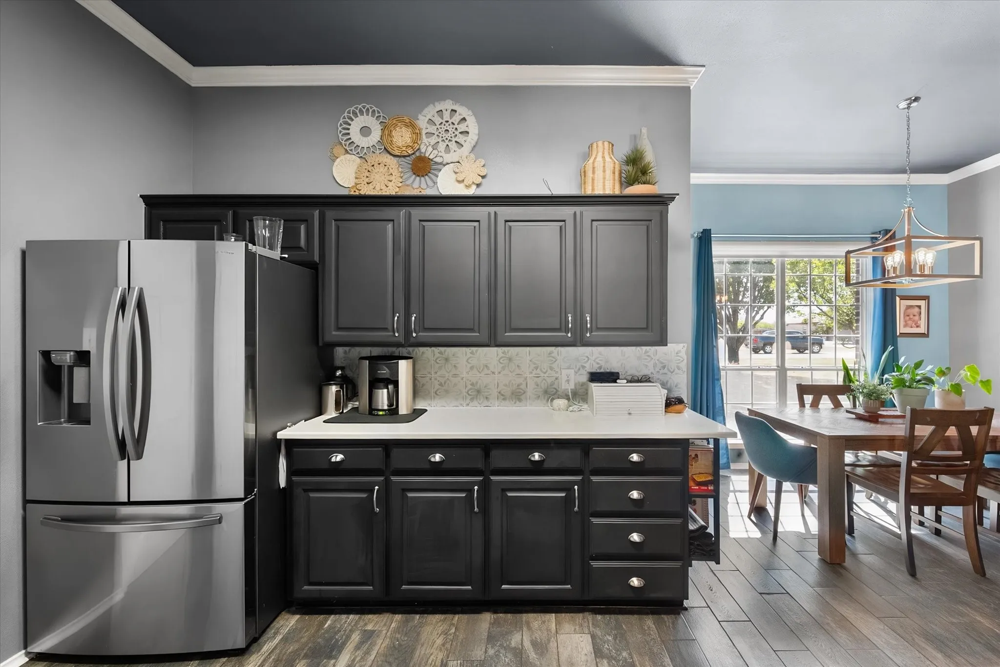 Stainless steel refrigerator, dark wood-finish flooring, white countertop, dark cabinetry with silver hardware, and a dining area with a rectangular chandelier