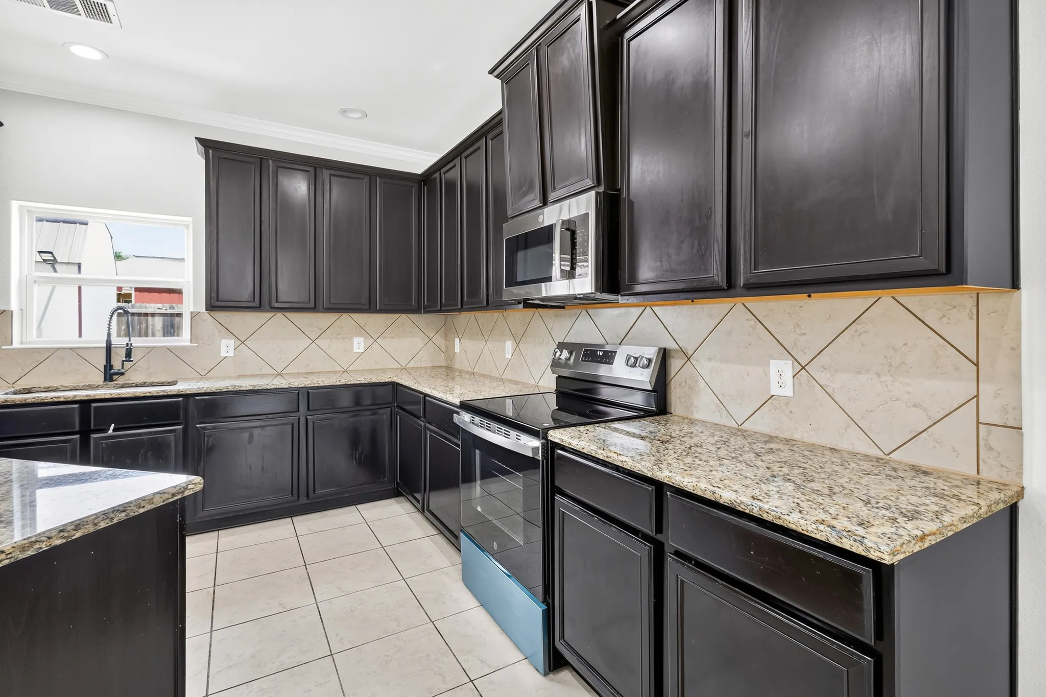 Kitchen featuring dark wood cabinetry, light-toned granite countertops, a diagonal tile backsplash, stainless steel appliances, and tile flooring
