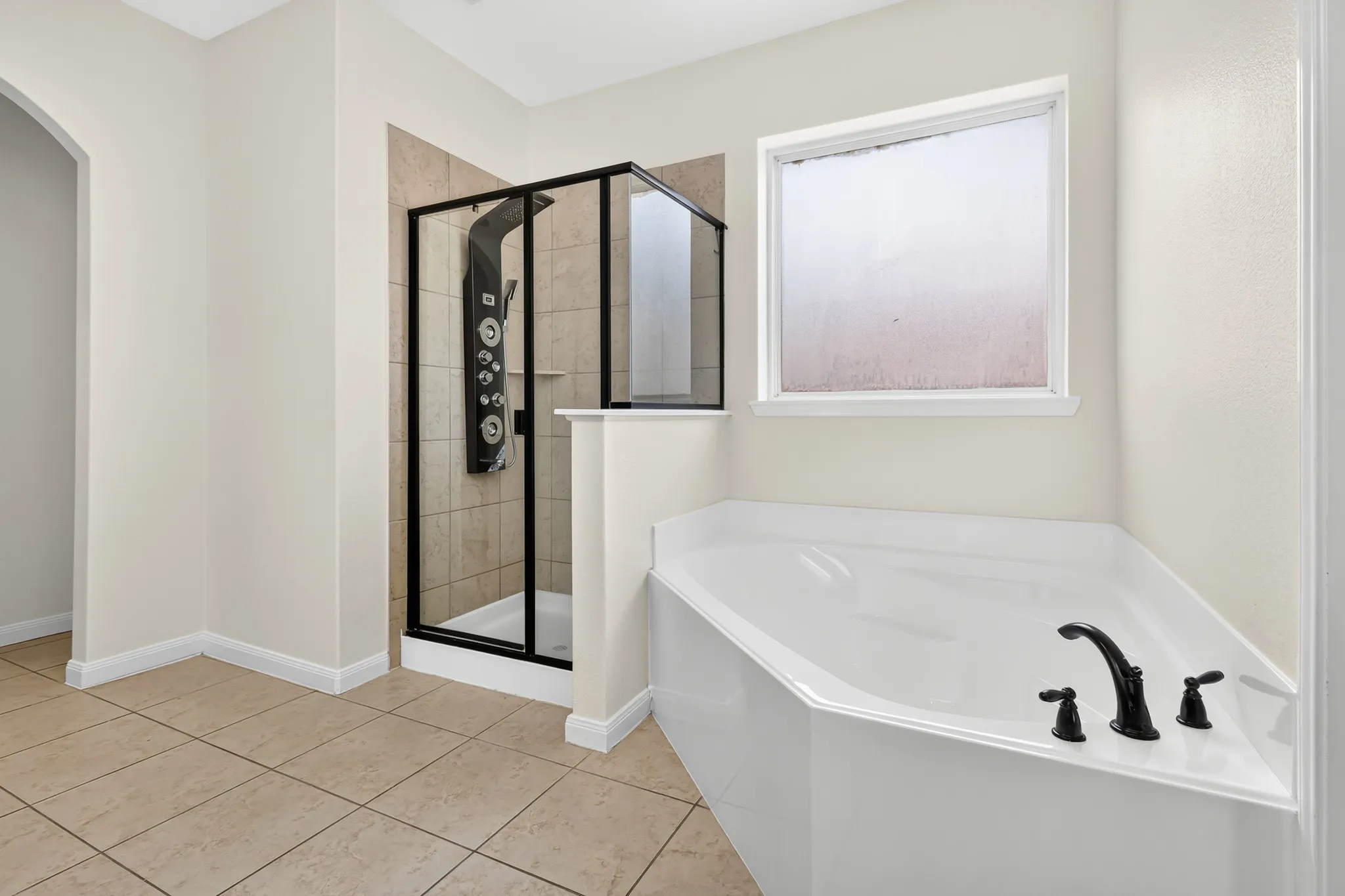 Tiled flooring, white soaking tub with dark-finish fixtures, and a glass-enclosed shower with a multi-function panel