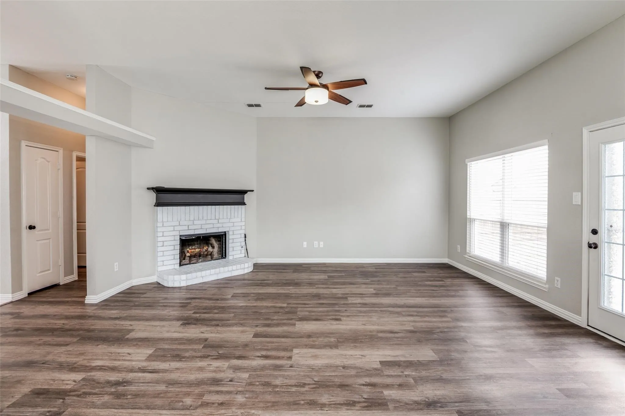 Spacious living area featuring wood-finish flooring, a white brick fireplace with a dark mantel, a ceiling fan, and multiple windows