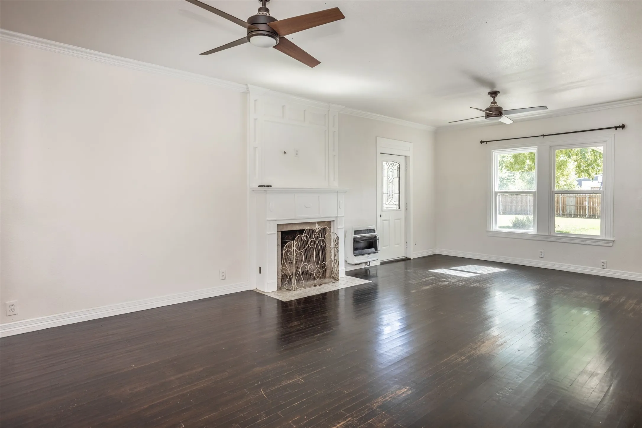 Dark wood-finish flooring, white fireplace with decorative screen, dual ceiling fans, and multiple windows