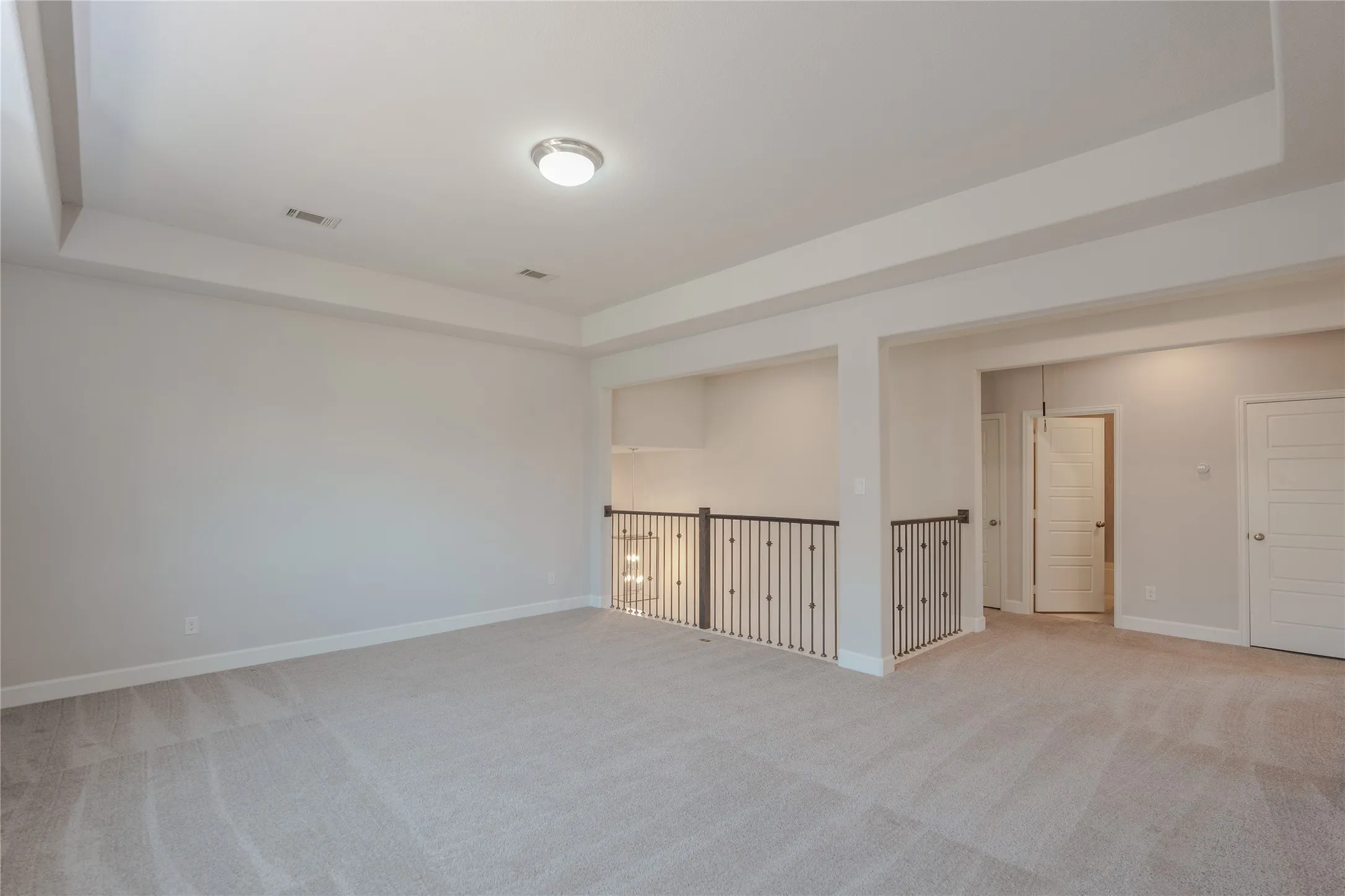 Spacious room featuring tray ceiling, flush mount lighting, light gray carpeting, and wrought iron railing