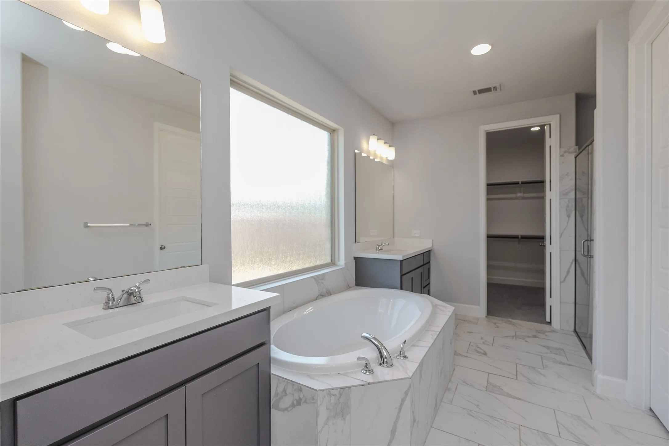 Spa-inspired primary bathroom featuring a freestanding soaking tub, dual-sink vanity with dark cabinetry and marble-finish countertops, large framed mirror with integrated lighting, walk-in closet, and tile flooring throughout