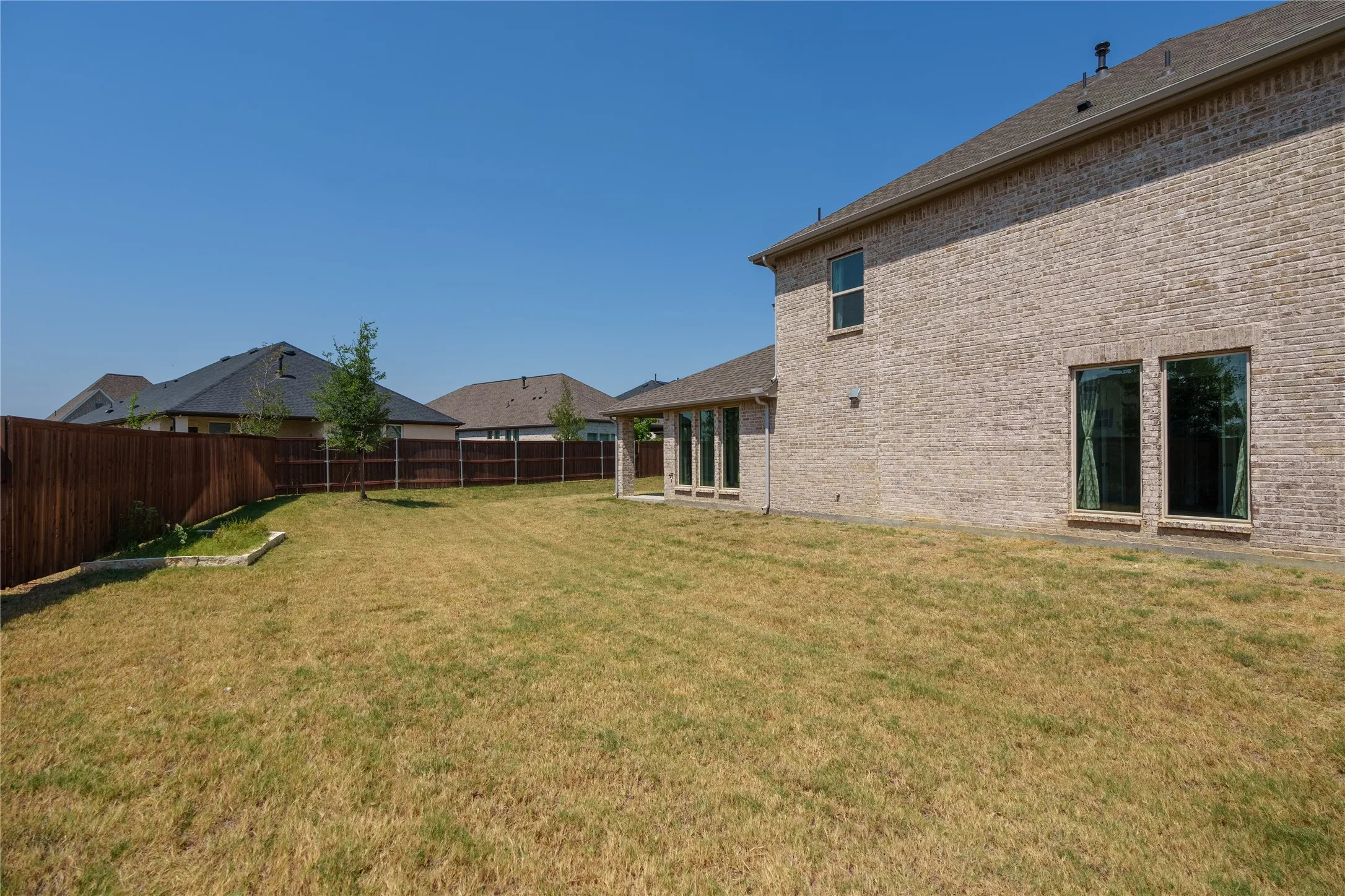 Expansive backyard with a full privacy fence, brick exterior, multiple windows, and a covered patio area