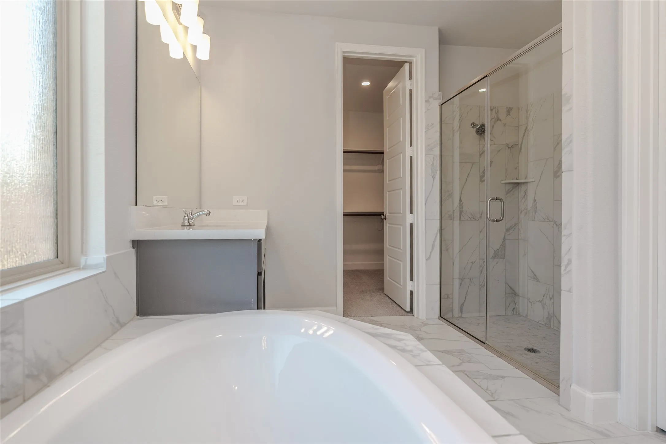 Soaking tub with surrounding white-veined tile, glass-enclosed shower with white-veined tile, single vanity with integrated sink and chrome faucet, and a walk-in closet with shelving