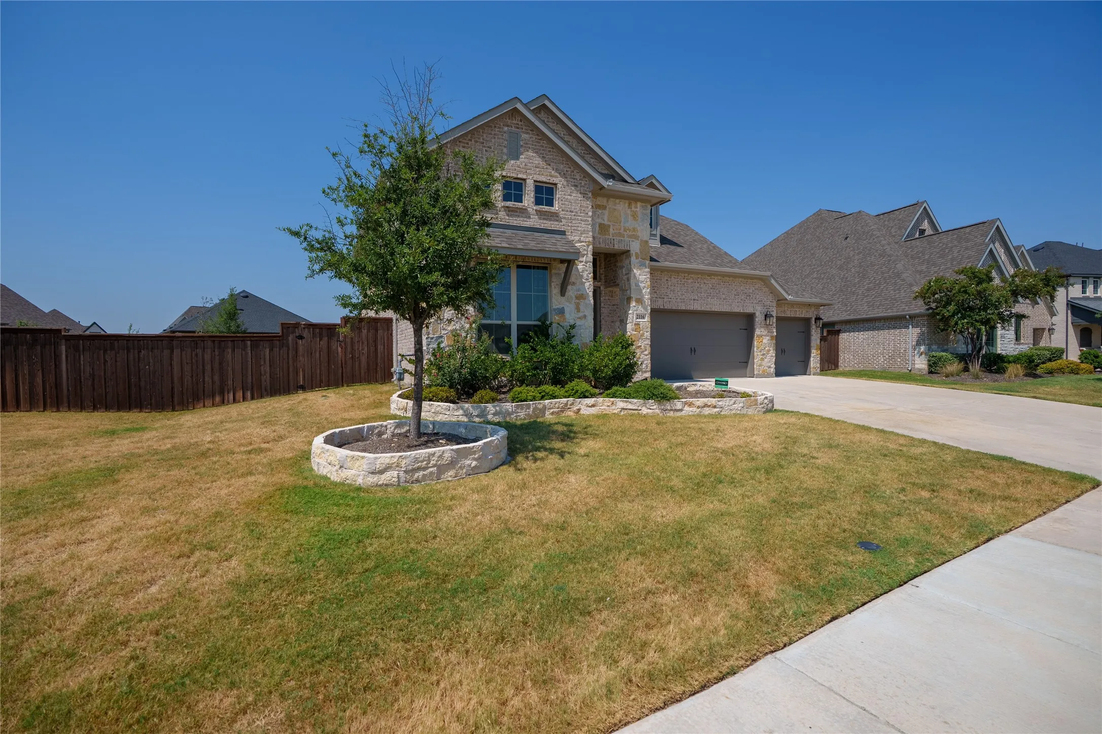 Stone and brick exterior with a multi-car garage, concrete driveway, and a fenced backyard