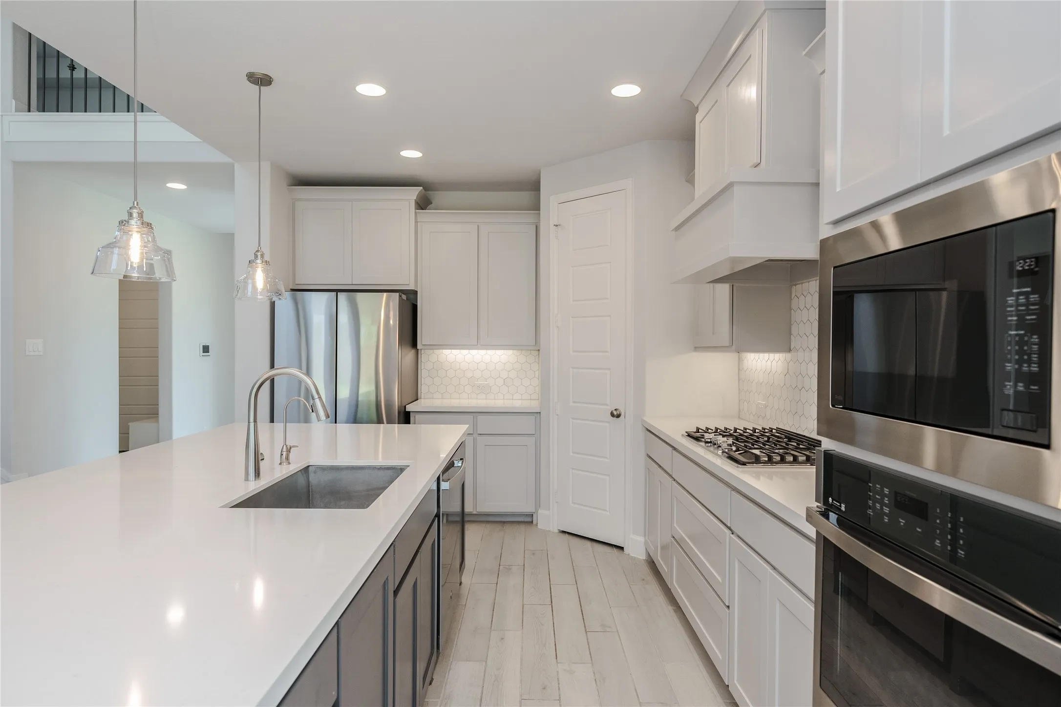 Spacious kitchen featuring a large island with an undermount sink, stainless steel appliances, white cabinetry, a hexagonal tile backsplash, and light wood-finish flooring
