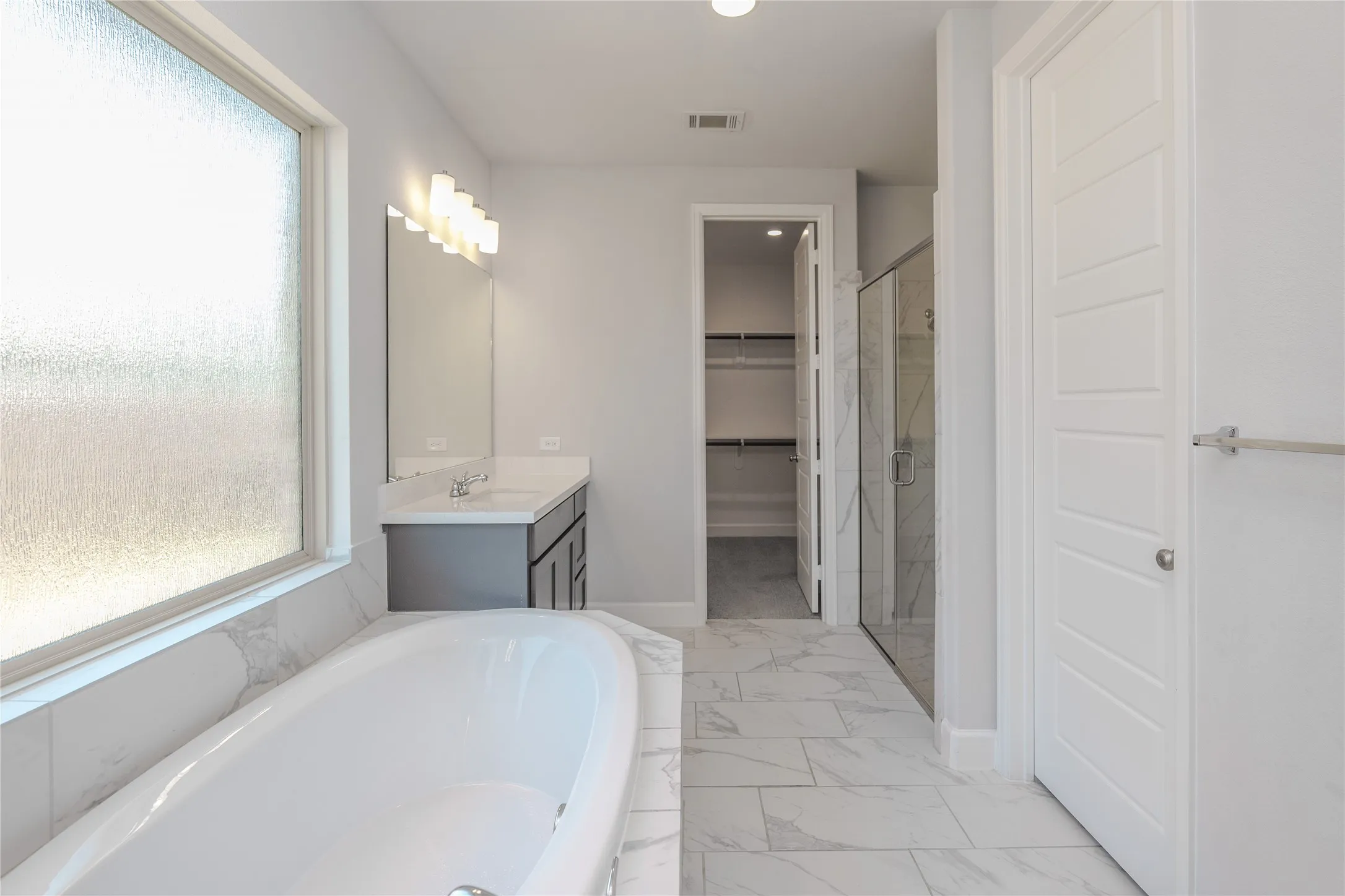 Freestanding soaking tub, double vanity with gray cabinetry, expansive walk-in shower with glass enclosure, large walk-in closet, and light-toned tile flooring
