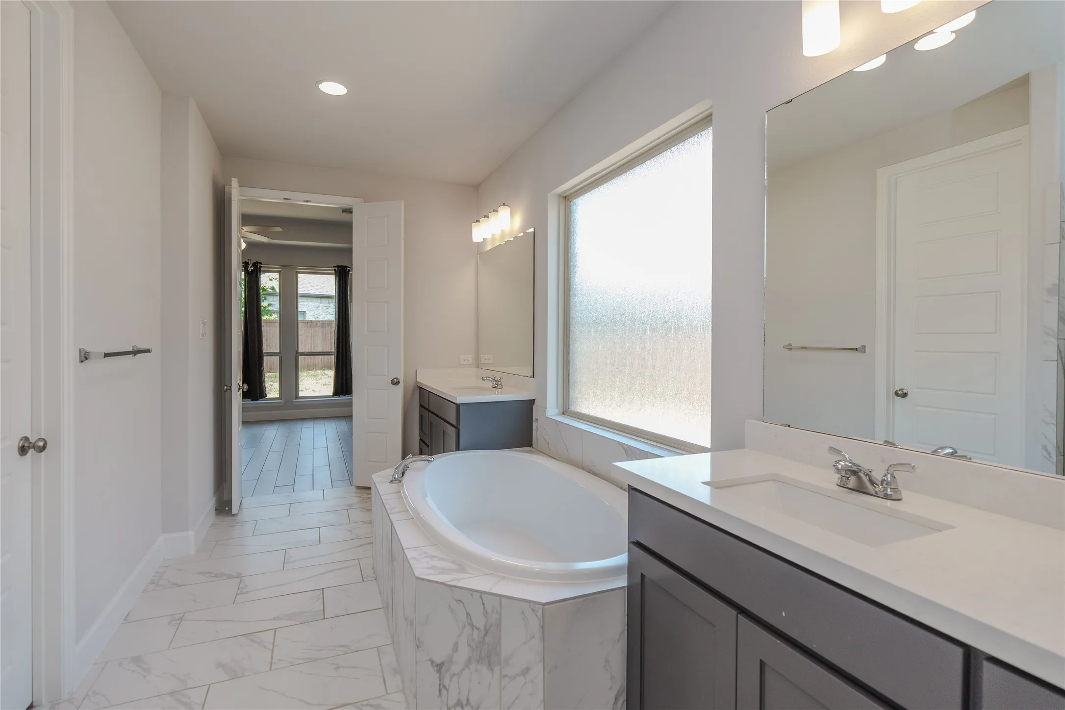Spa-inspired primary bathroom featuring a freestanding soaking tub positioned beneath an expansive window, dual vanity with quartz countertop and undermount sinks, large framed mirror, recessed ceiling lighting, and herringbone-pattern tile flooring
