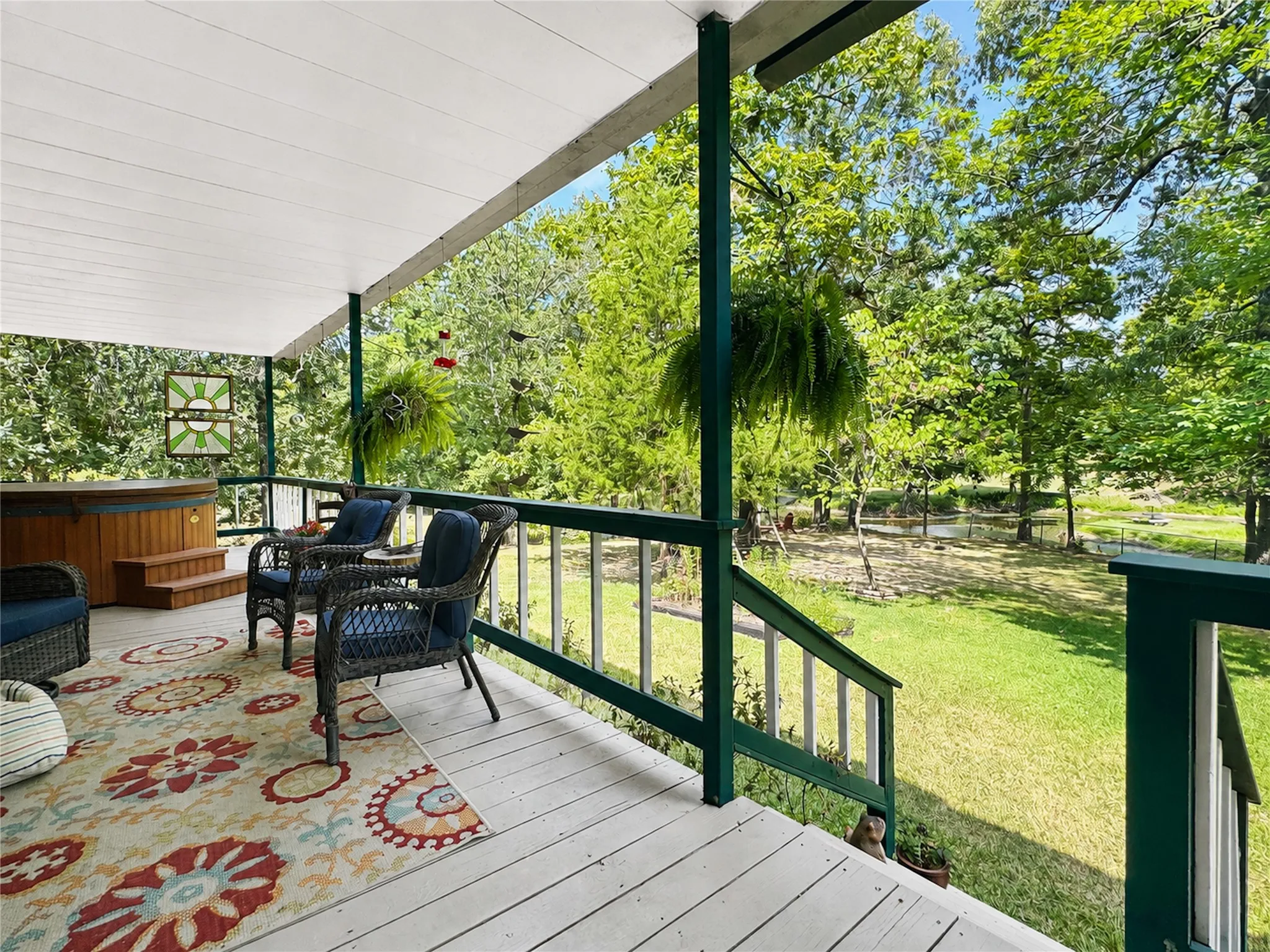 Covered deck with a hot tub, painted wood railing, and plank flooring overlooking a landscaped yard with mature trees