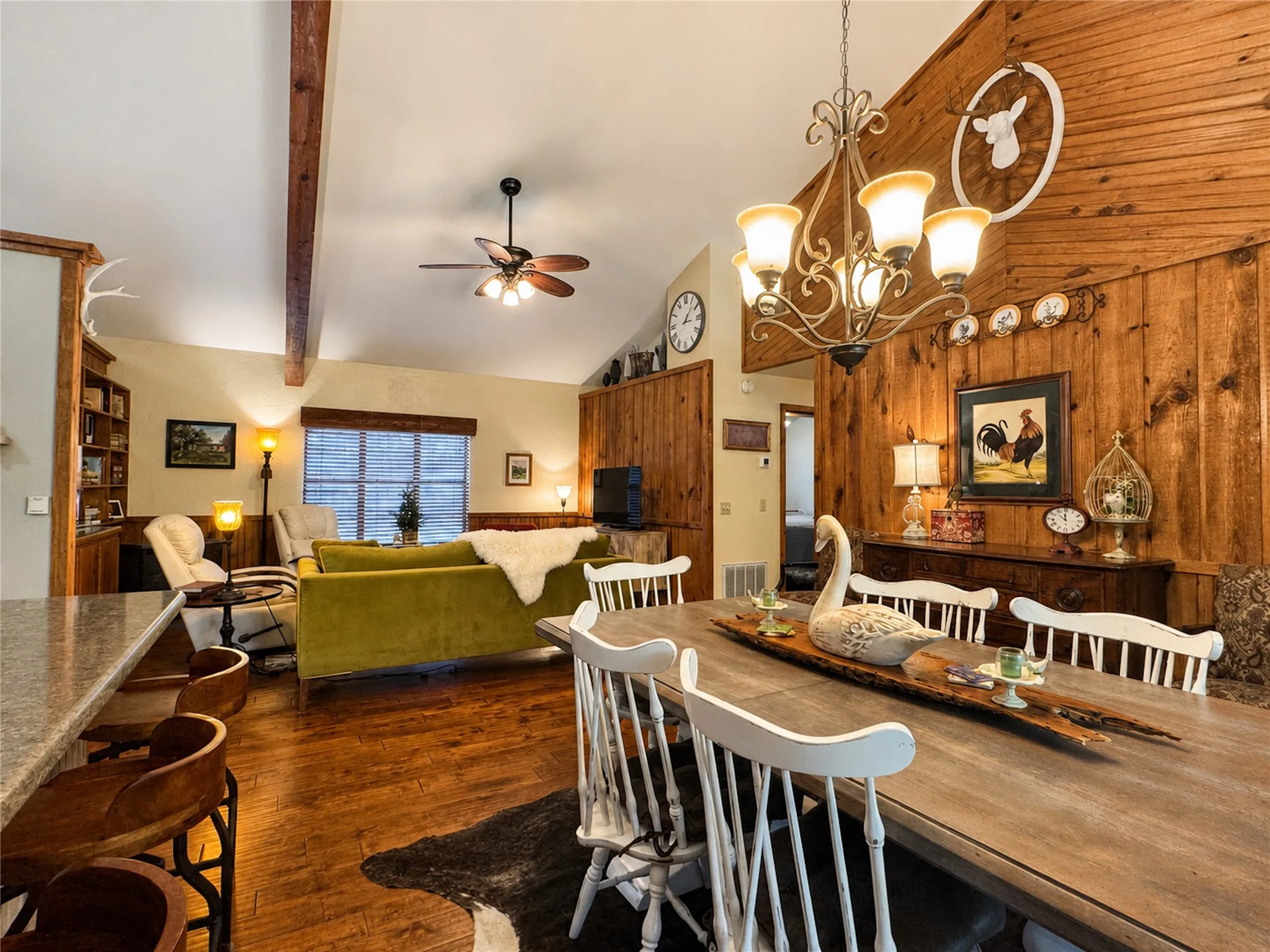 Vaulted ceiling with exposed wood beams, wood-finish flooring, wood panel accent wall, ceiling fan, and built-in shelving