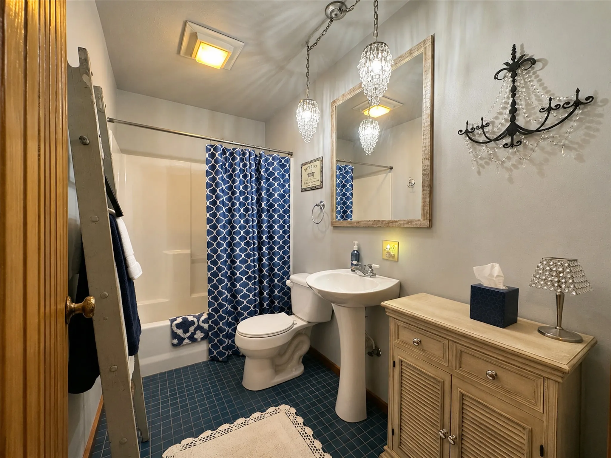 Pedestal sink with chrome faucet, bath/shower combination with white surround, blue grid-patterned flooring, and a mirrored vanity with wood-finish frame