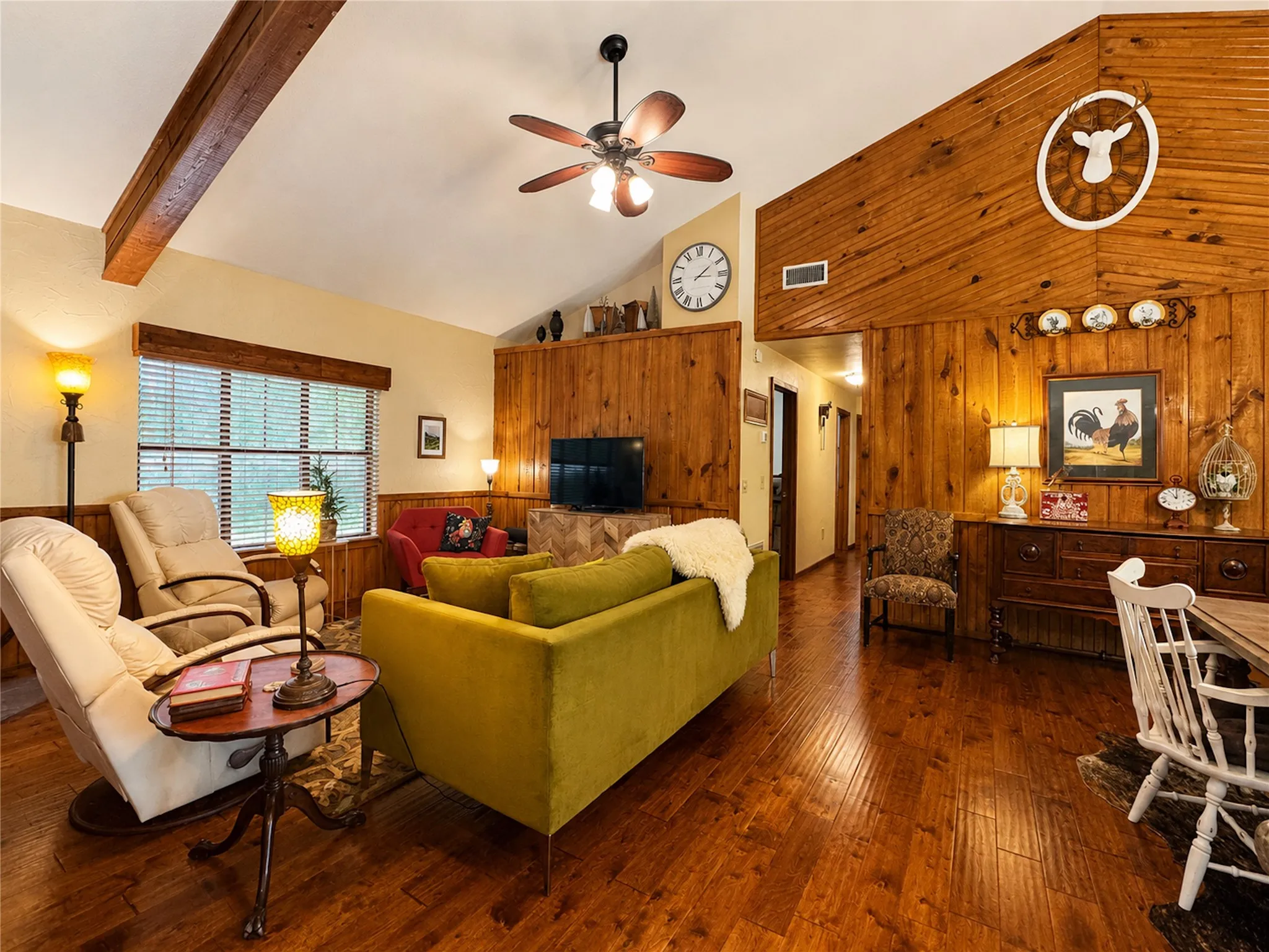 Vaulted ceiling with wood beam accents, wood-finish flooring, wood paneling, ceiling fan with light fixture, and large window with blinds