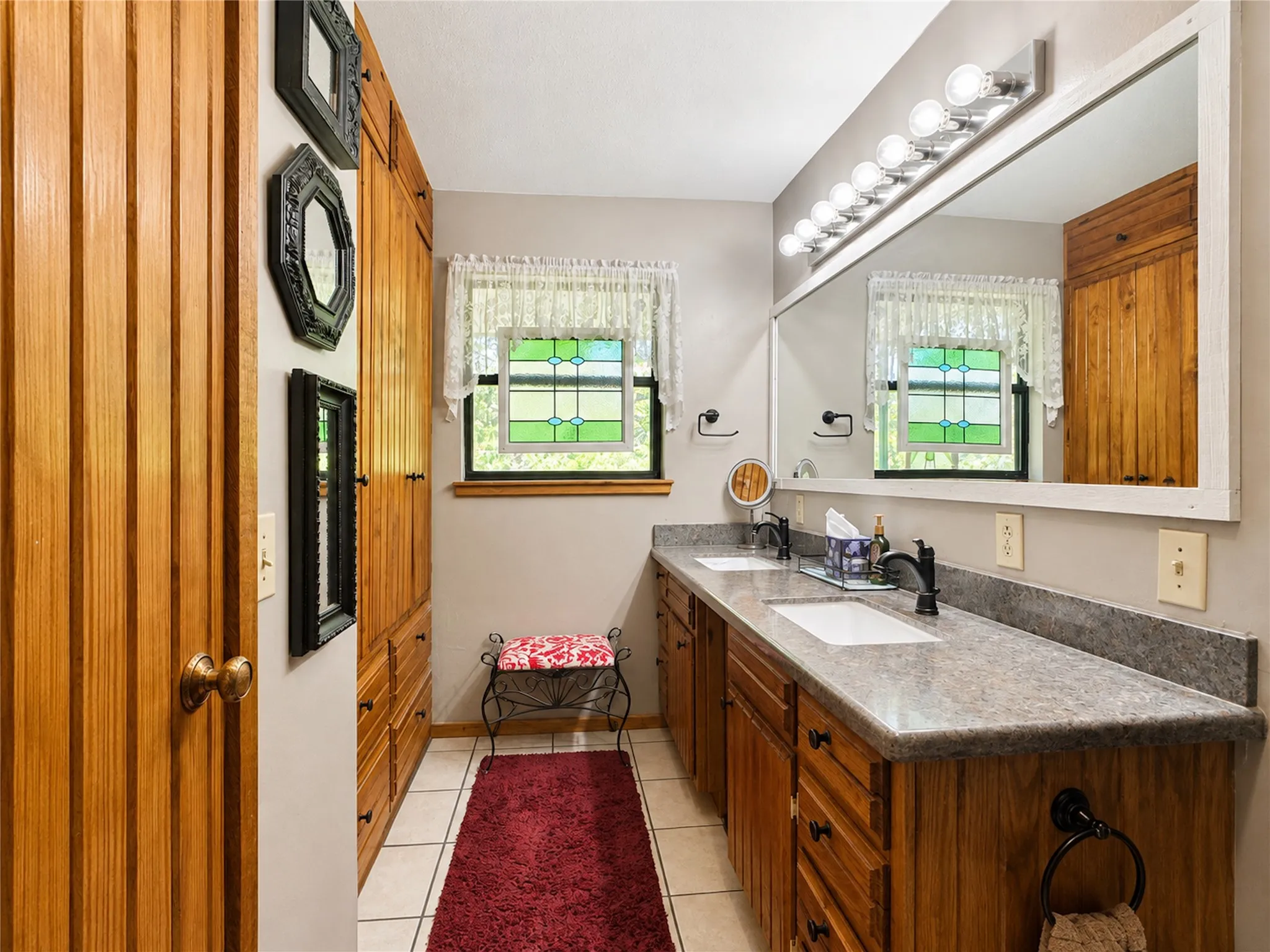 Elegant primary bathroom featuring warm wood cabinetry with ample storage, dual mirrors with integrated lighting, polished stone countertops, and ceramic tile flooring