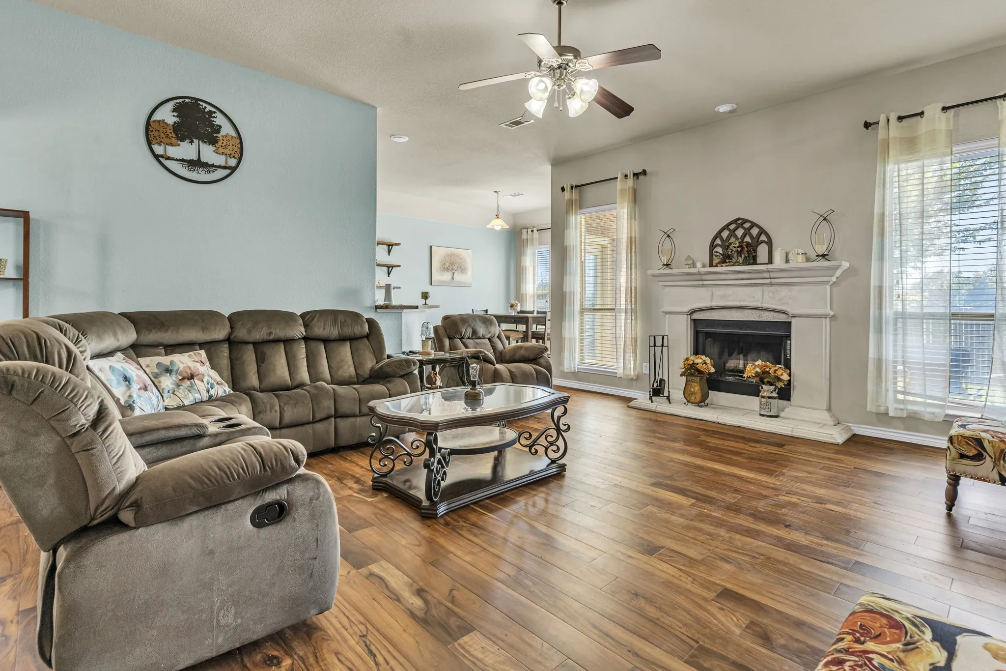 Wood-finish flooring throughout, a white stone fireplace with a substantial mantel, and multiple windows allowing natural light