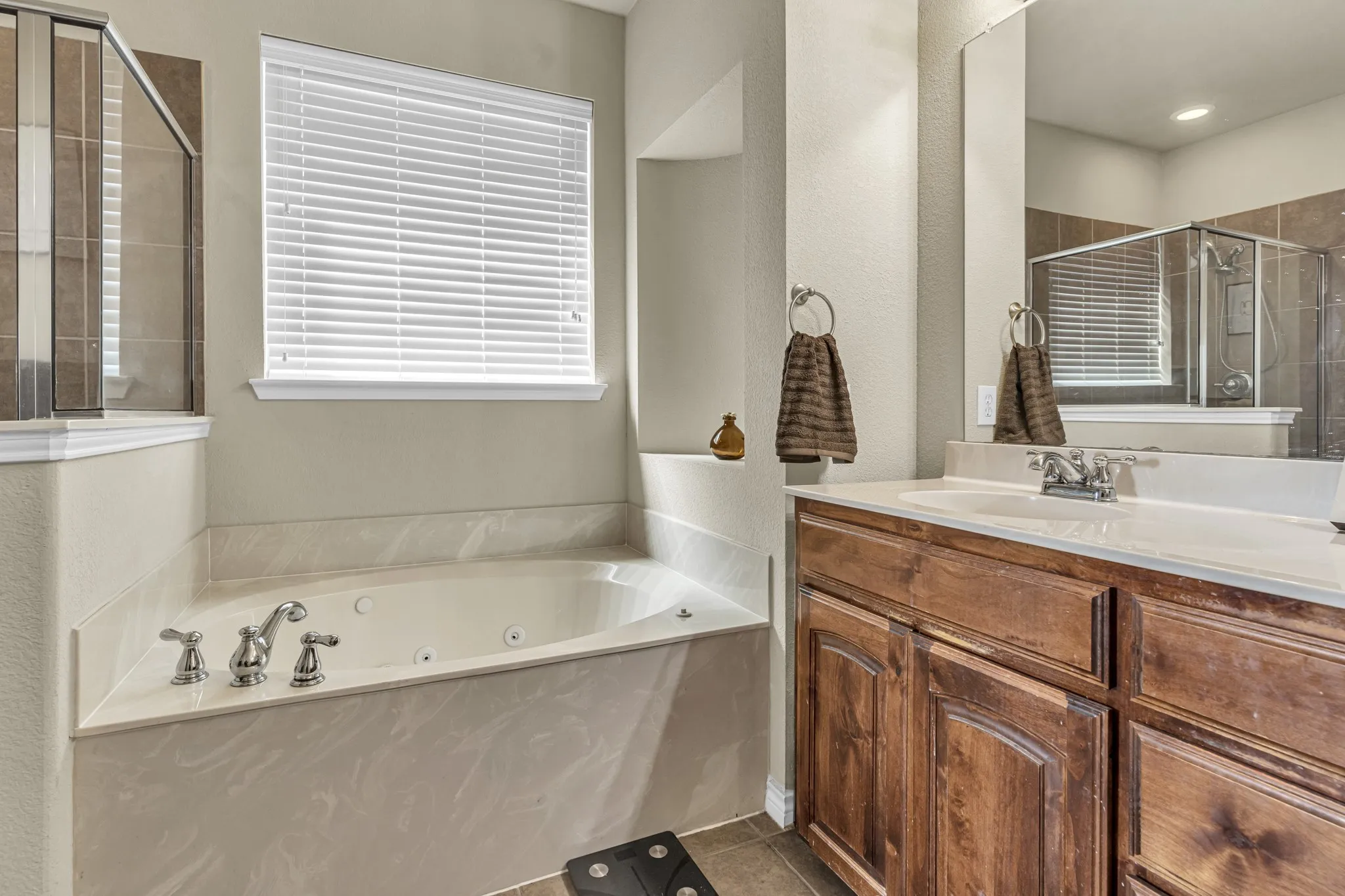 Built-in jetted tub, wood-finish vanity with ample storage, integrated white sink, expansive wall mirror, and a glass-enclosed shower with tiled walls