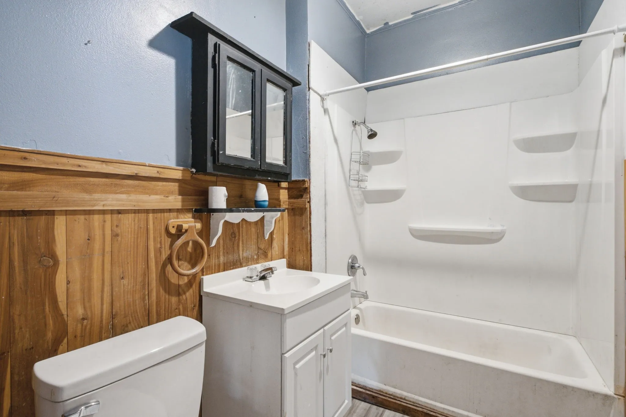Wall-mounted black medicine cabinet, wood-finish wainscoting, white vanity with integrated sink, white toilet, and a white shower-over-tub unit