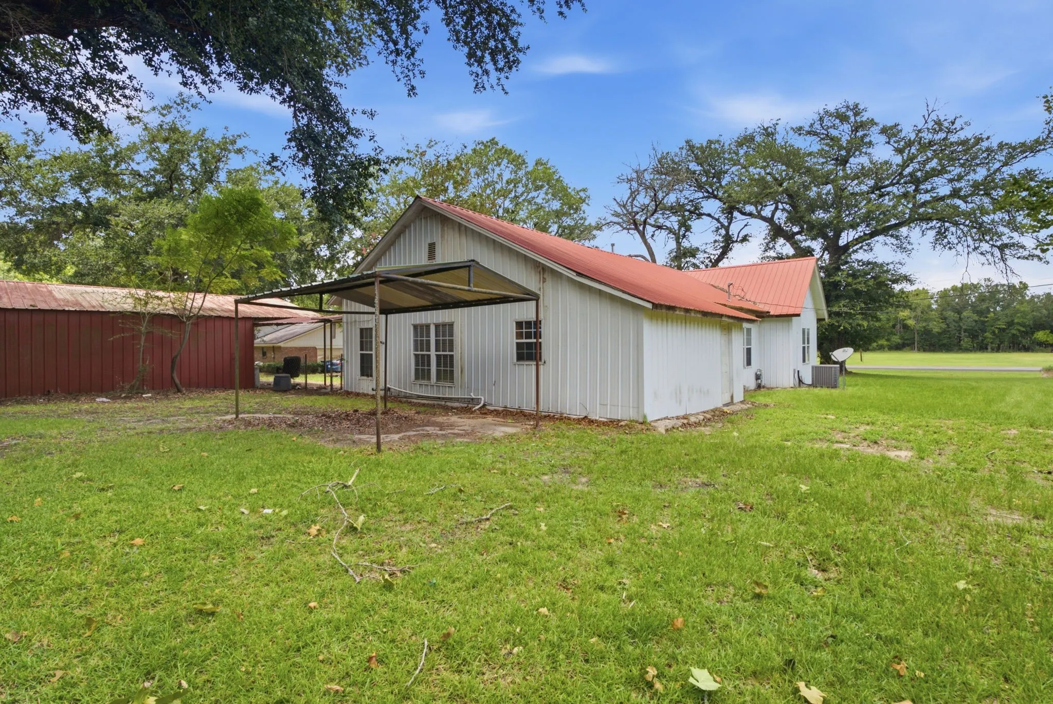 Expansive grassy yard with mature trees, a red metal roof, white siding, and a covered patio structure