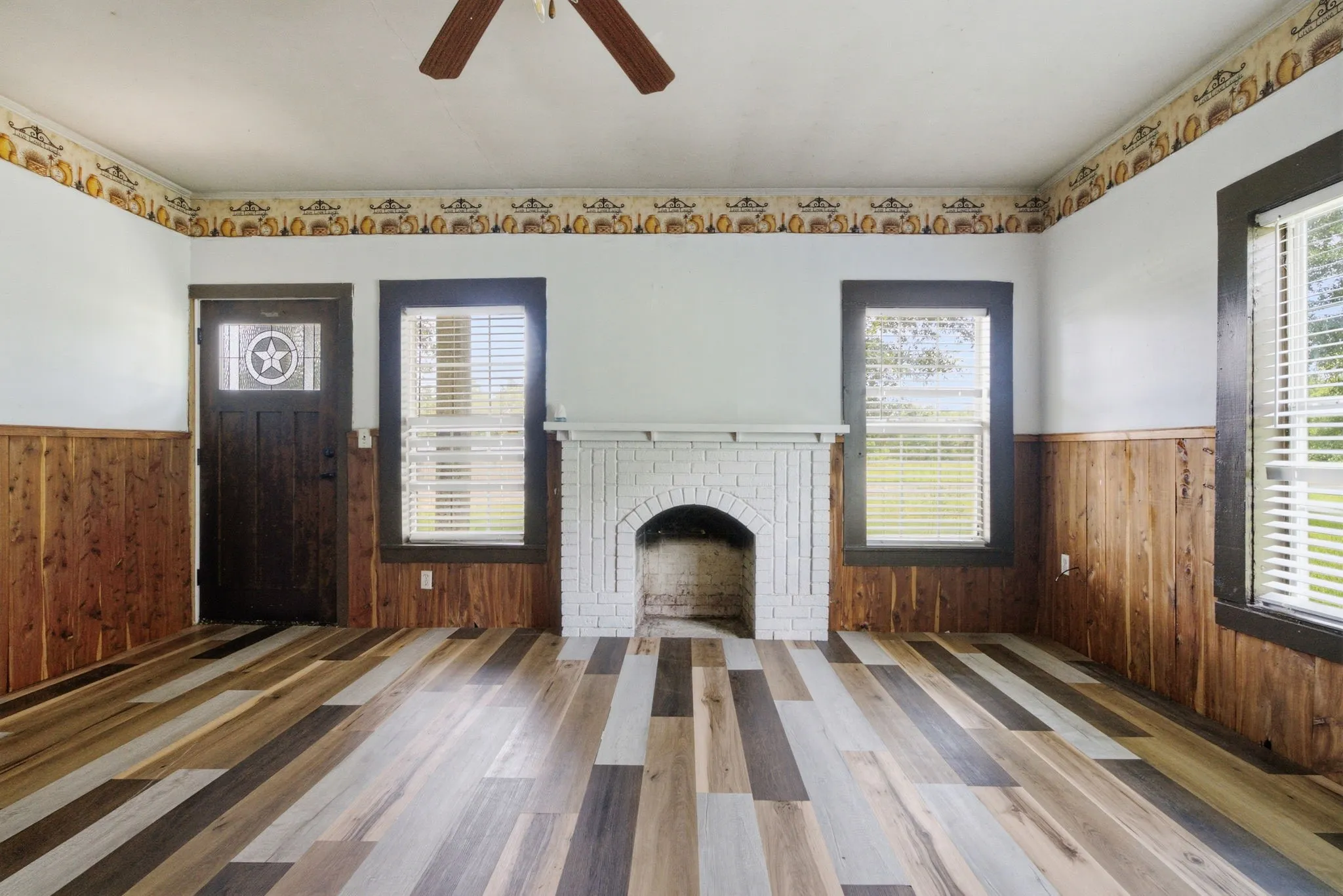Multi-tone wood-finish flooring, white brick fireplace, wood wainscoting, and a ceiling fan