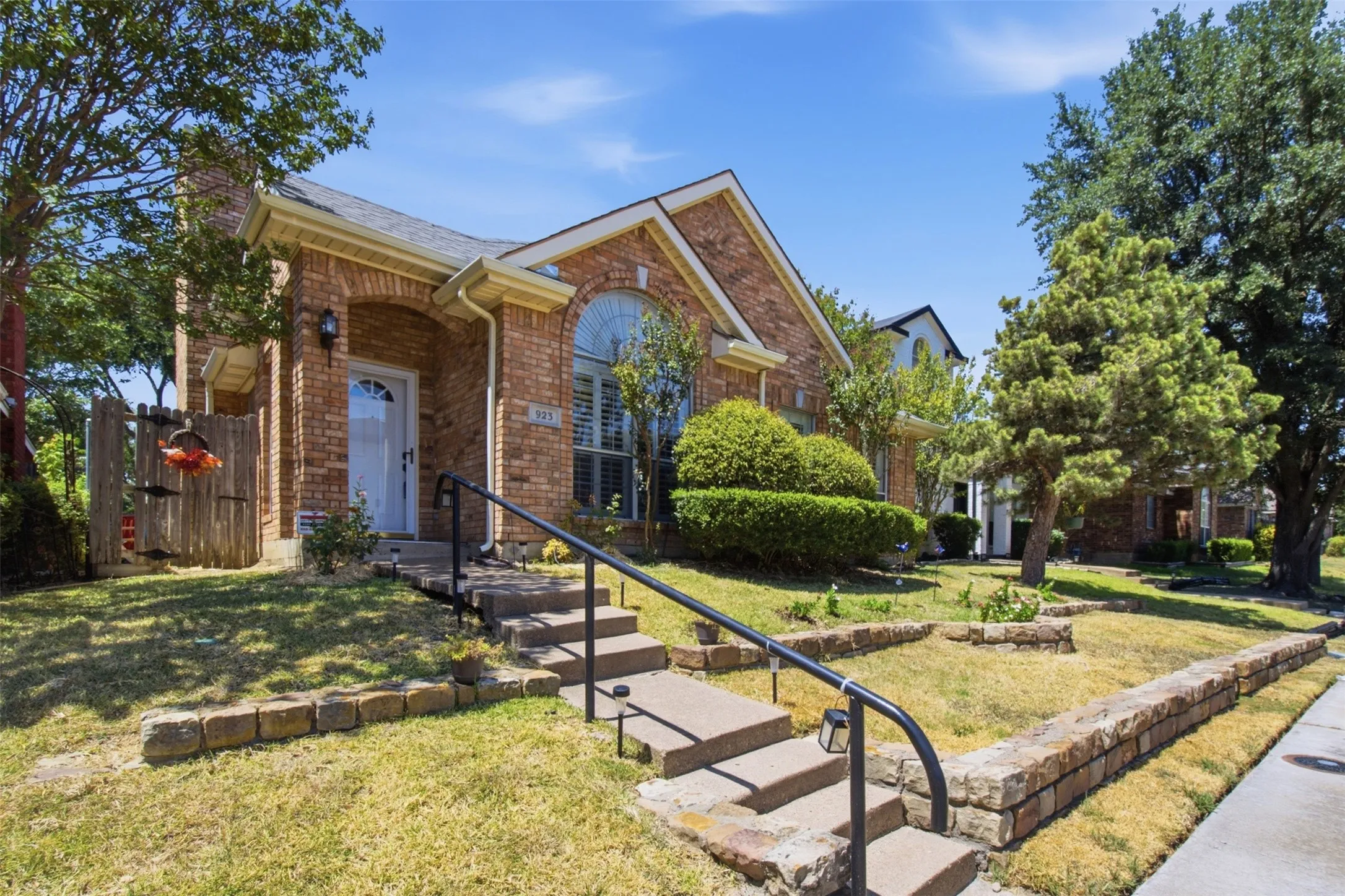 Brick exterior with arched entryway, front porch with black railing, arched window with interior shutters, and mature landscaping