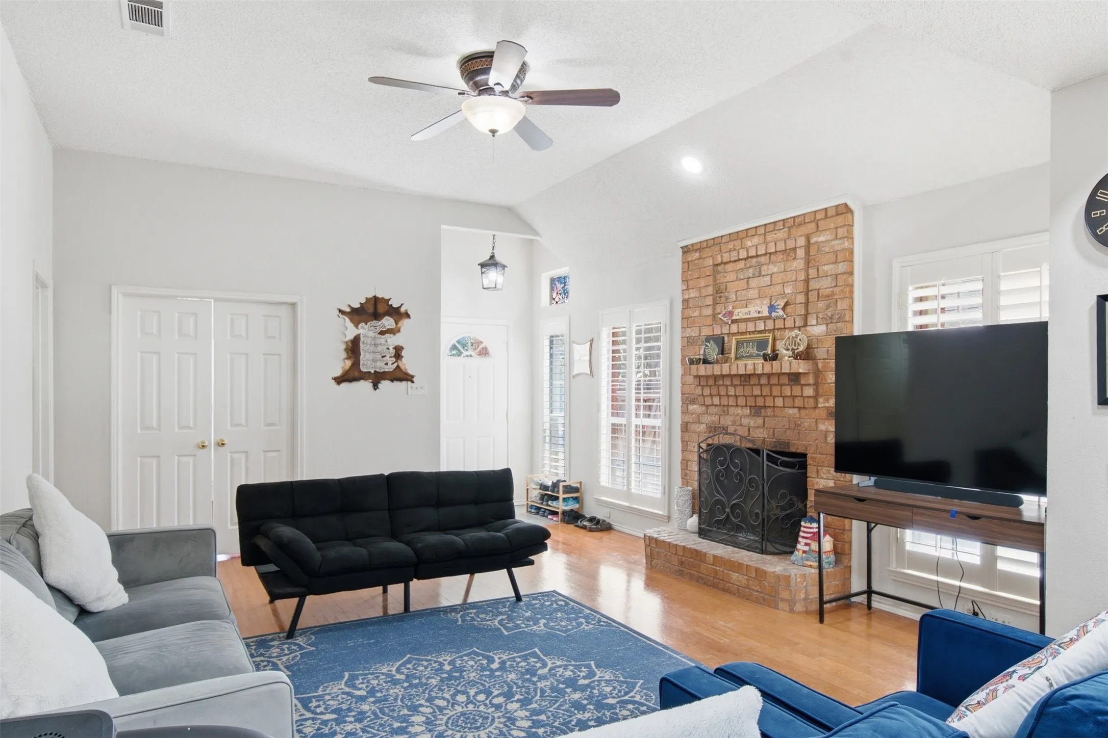 Vaulted ceiling with a ceiling fan, brick fireplace, wood-finish flooring, and white plantation shutters